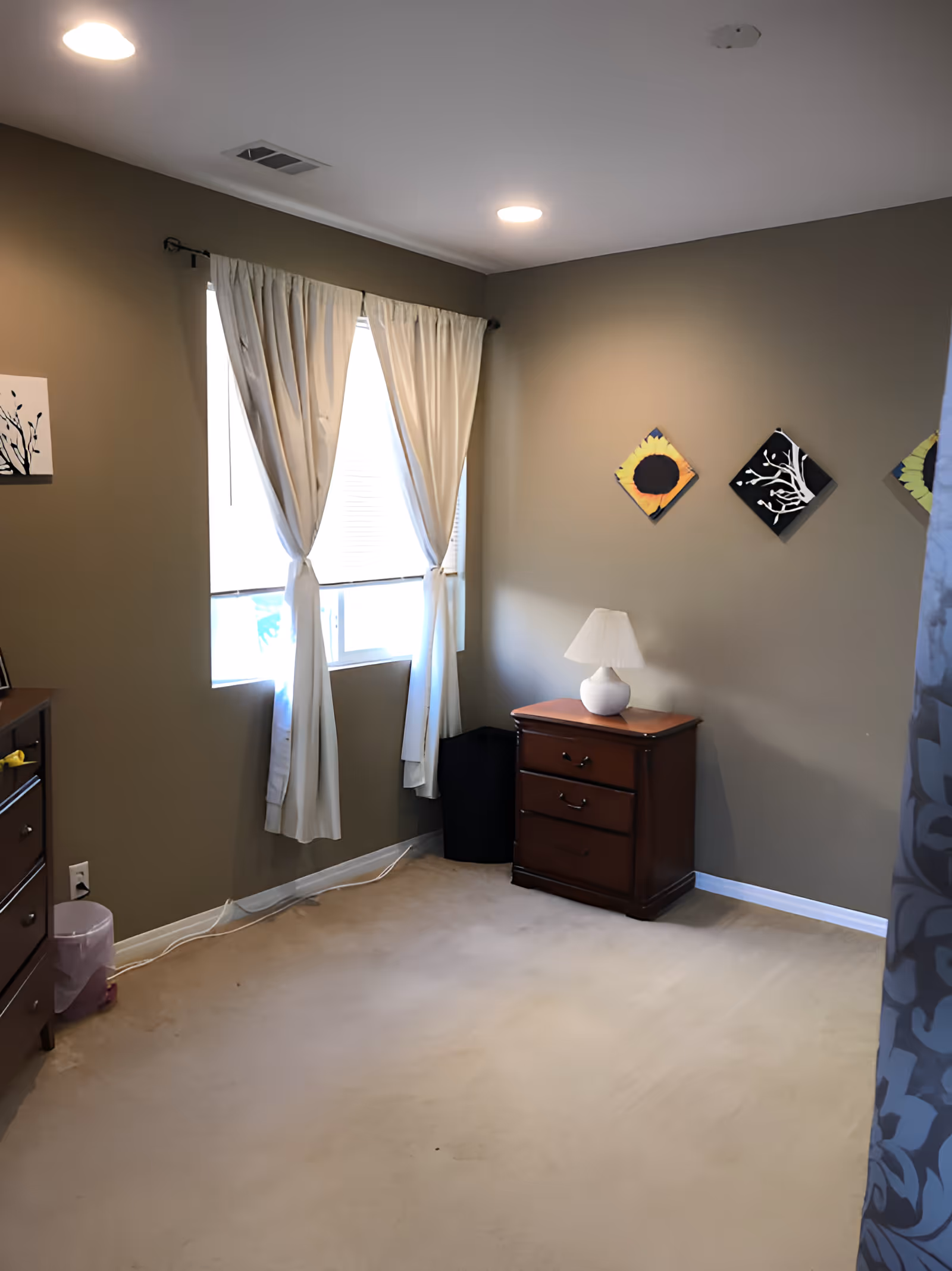 Small bedroom with a window and white curtains, a wooden nightstand with a lamp, dresser, and wall art.