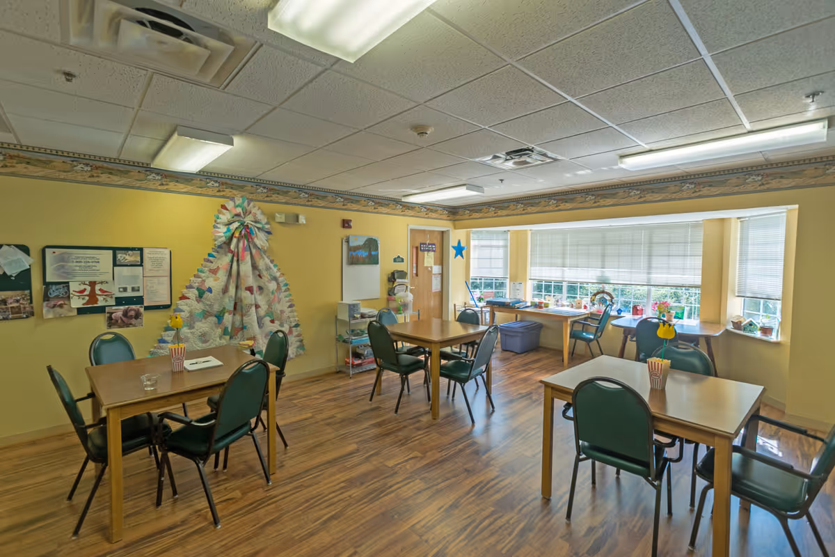 Communal dining/activity room with several wooden tables, green chairs, craft supplies and large windows letting in daylight.