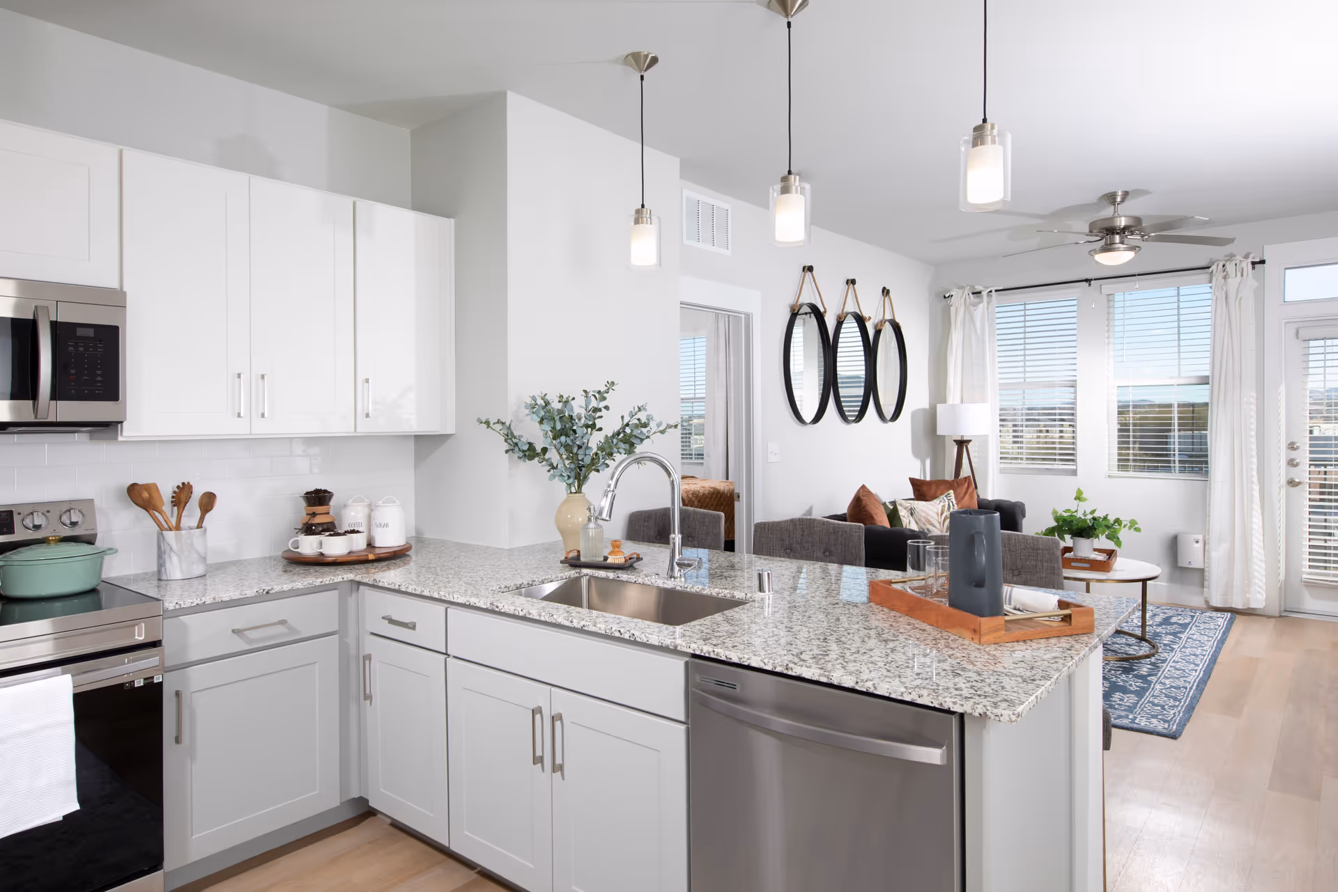 Modern kitchen with white and gray cabinets, granite countertops, stainless steel appliances including a microwave, stove, and dishwasher. The kitchen island has a sink and a decorative tray with a pitcher and glasses. In the background, there is a living room area with a sofa, pillows, a floor lamp, three round mirrors on the wall, and large windows with white curtains letting in natural light.