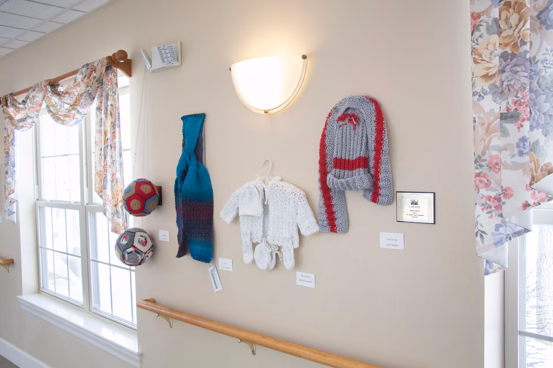 A hallway wall decorated with handmade knitted items including scarves, baby clothes, and hats, displayed under a wall light. Two large windows with floral curtains allow natural light into the space. A wooden handrail runs along the wall beneath the display.
