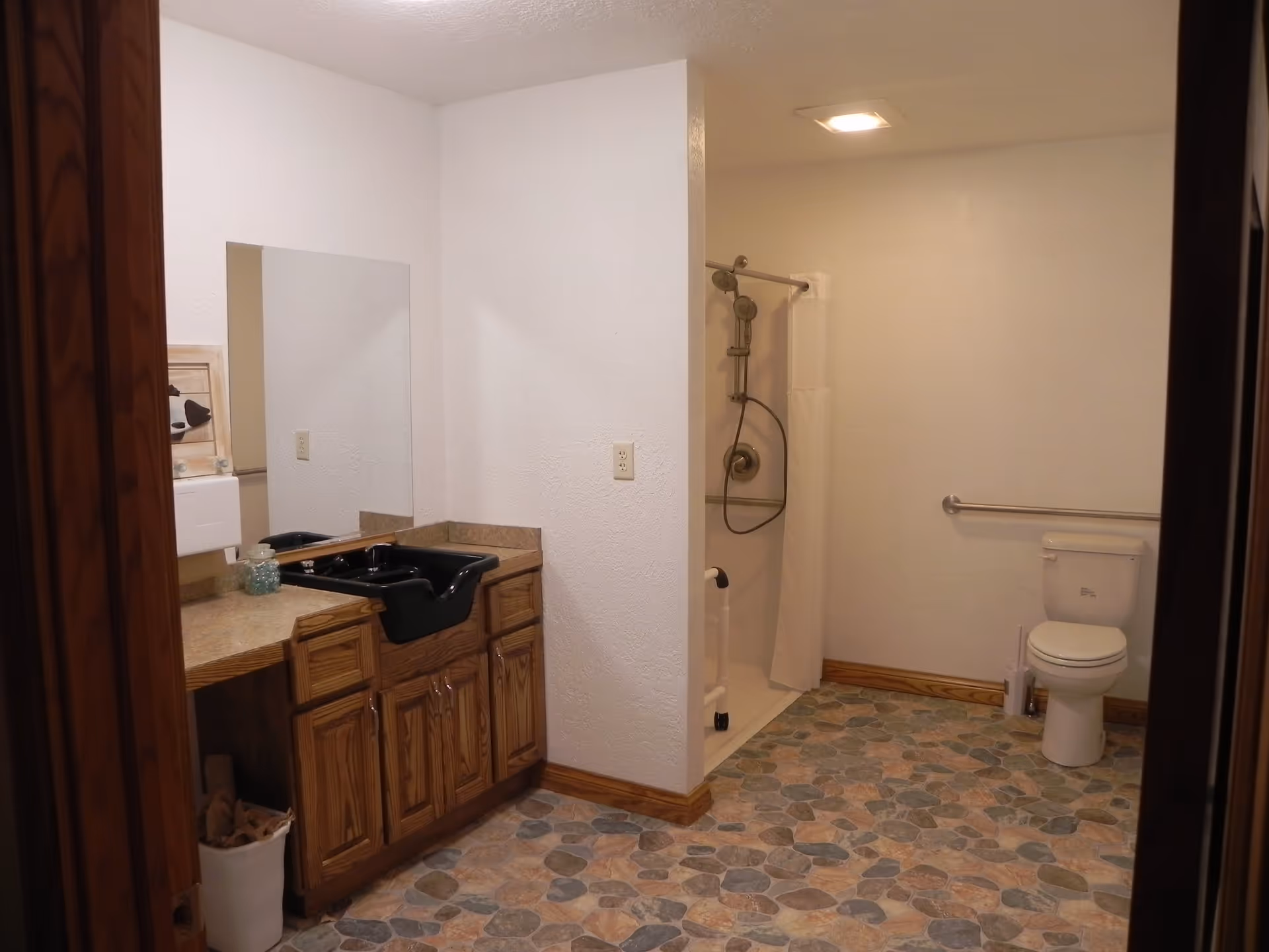 A bathroom with a black sink set in a wooden cabinet with a large mirror above it. To the right, there is a walk-in shower with a grab bar and a white shower curtain. Further right, there is a toilet with a grab bar on the wall behind it. The floor has a stone-patterned tile, and the walls are painted white.