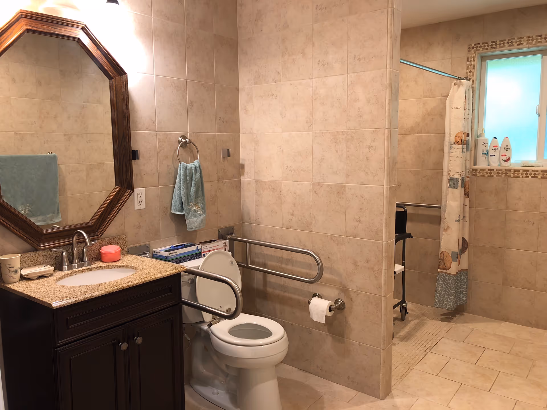 A spacious bathroom with beige tiled walls and floor. It features a dark wood vanity with a granite countertop and an octagonal mirror above it. There is a toilet with safety grab bars on both sides. A towel ring with a blue towel is mounted on the wall next to the toilet. In the background, there is a walk-in shower area with a curtain and a window above it. Several bottles of shampoo and body wash are placed on the window sill.