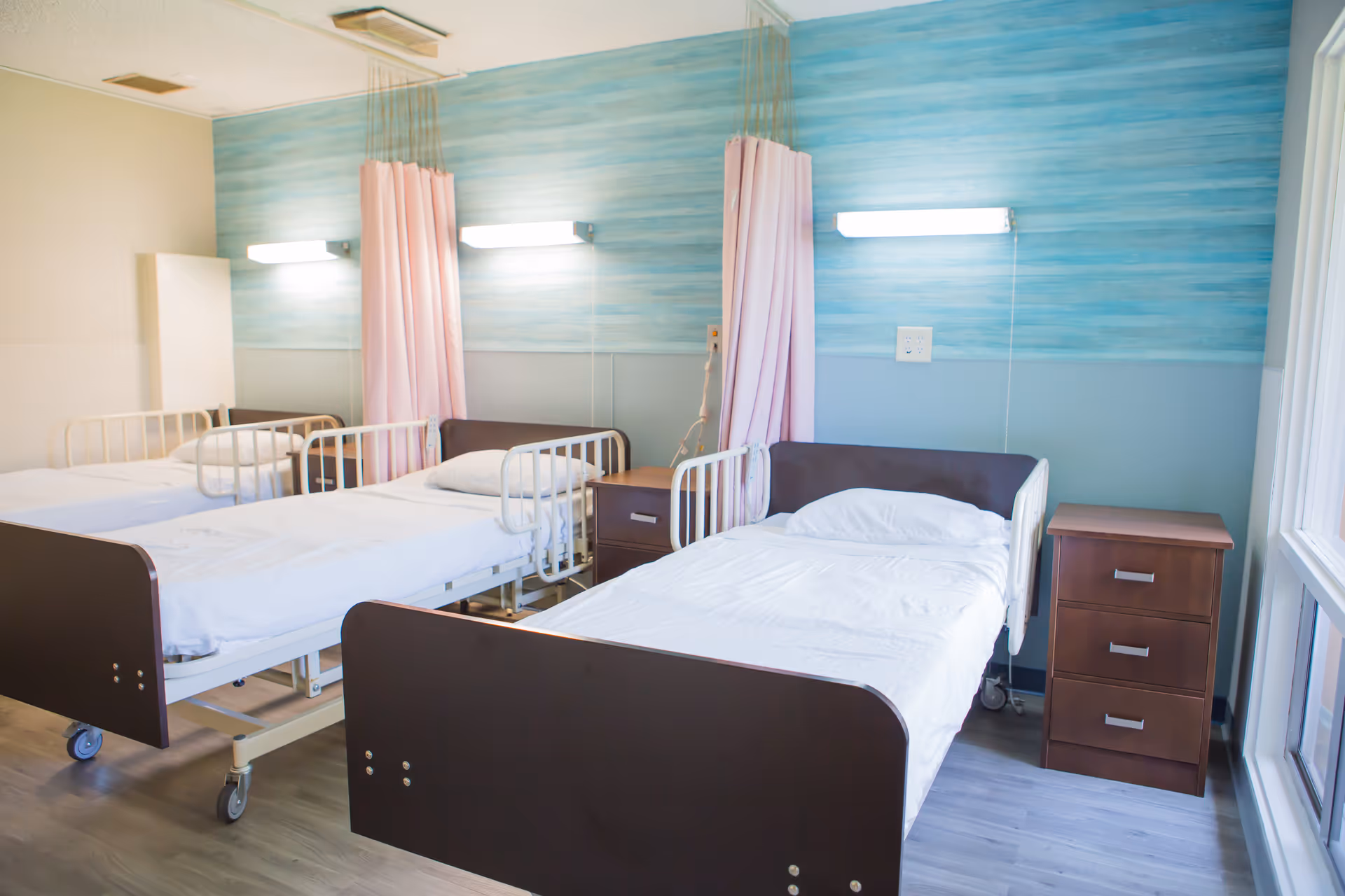 A clean and bright healthcare facility room with three hospital beds, each equipped with white bedding and pillows. The beds have side rails and are separated by pink privacy curtains. The walls have a blue and gray color scheme, and there are wooden bedside tables next to the beds. The room has large windows allowing natural light to enter.