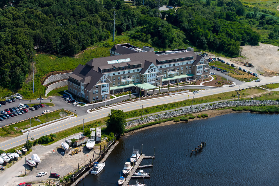 Aerial view of a multi-story senior living building on the waterfront with parking lots, a road, and boats nearby.
