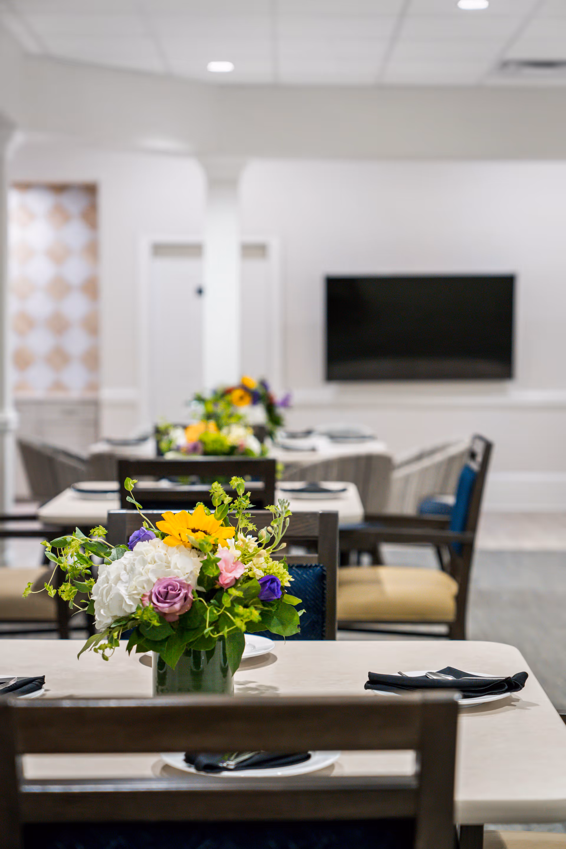 A dining area in a senior living facility with tables set with plates and black napkins. Each table has a floral centerpiece featuring sunflowers, white hydrangeas, purple and pink flowers, and greenery. Chairs with blue and beige cushions surround the tables. A flat-screen TV is mounted on the white wall in the background.