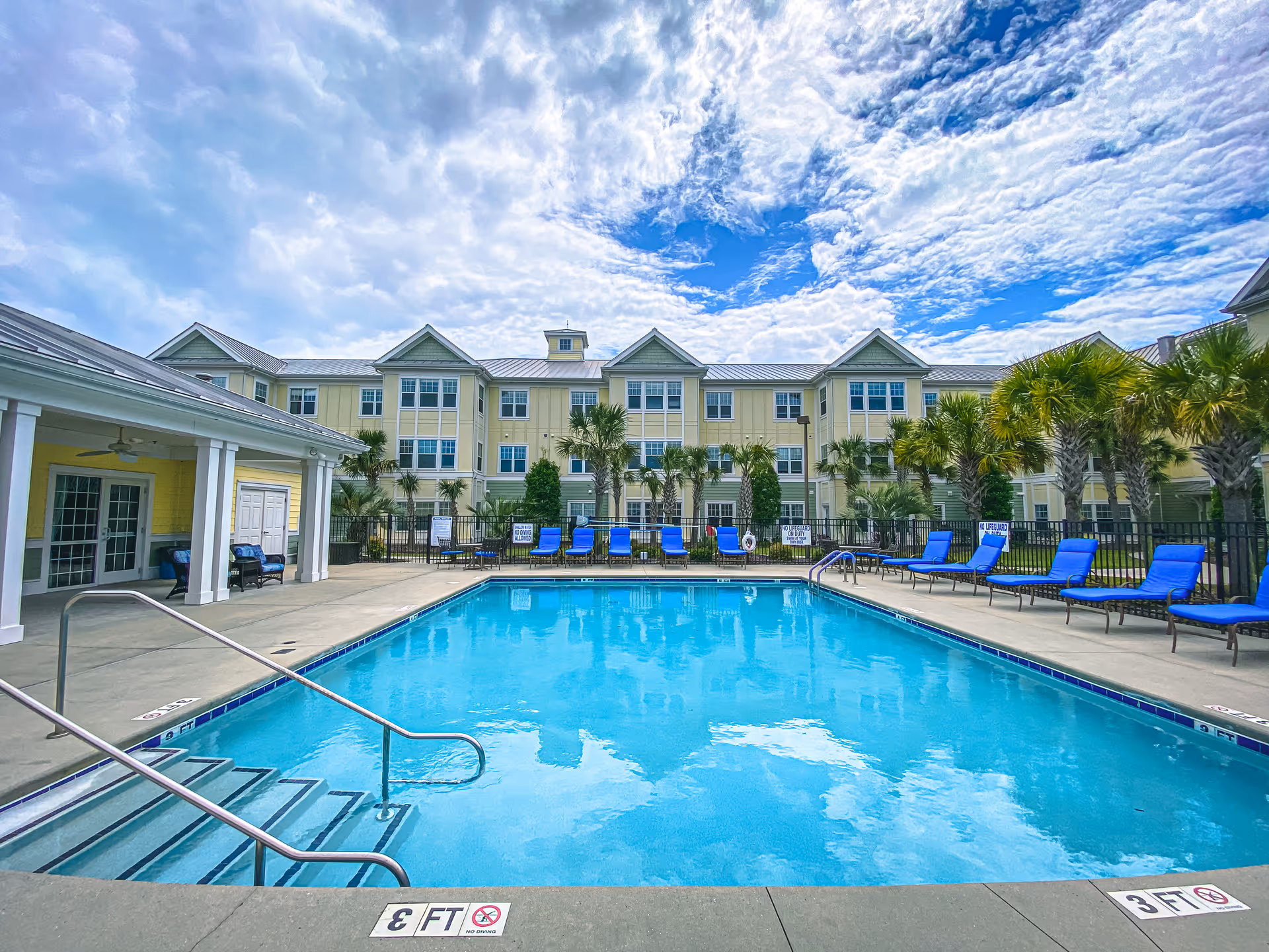Outdoor swimming pool with clear blue water surrounded by a concrete deck and blue lounge chairs. The pool area is fenced and palm trees line the perimeter. In the background, there is a three-story yellow building with white trim under a partly cloudy blue sky.