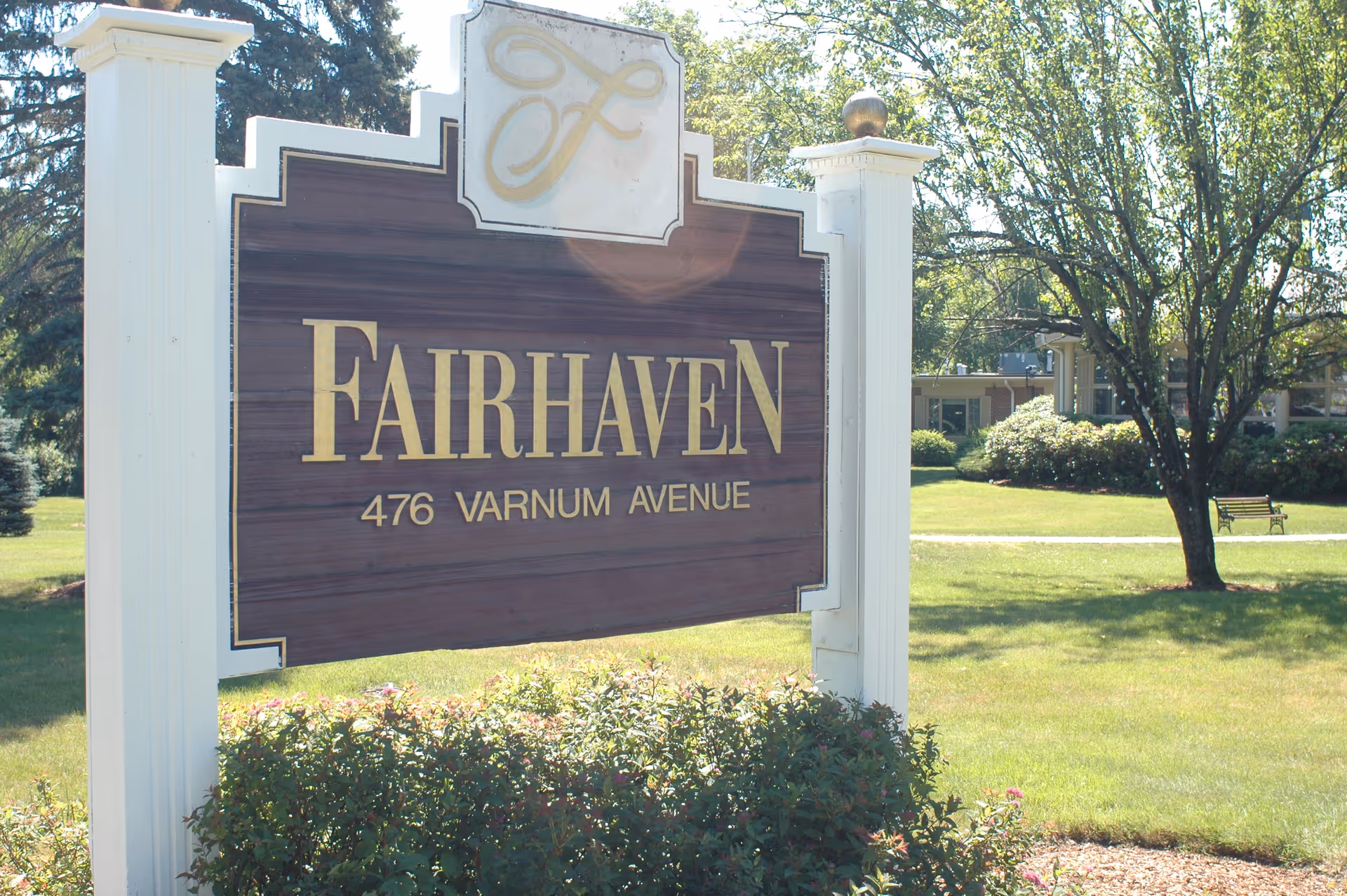 Outdoor view of a large wooden sign with white posts that reads 'Fairhaven 476 Varnum Avenue' surrounded by green grass, bushes, and trees with a building visible in the background.