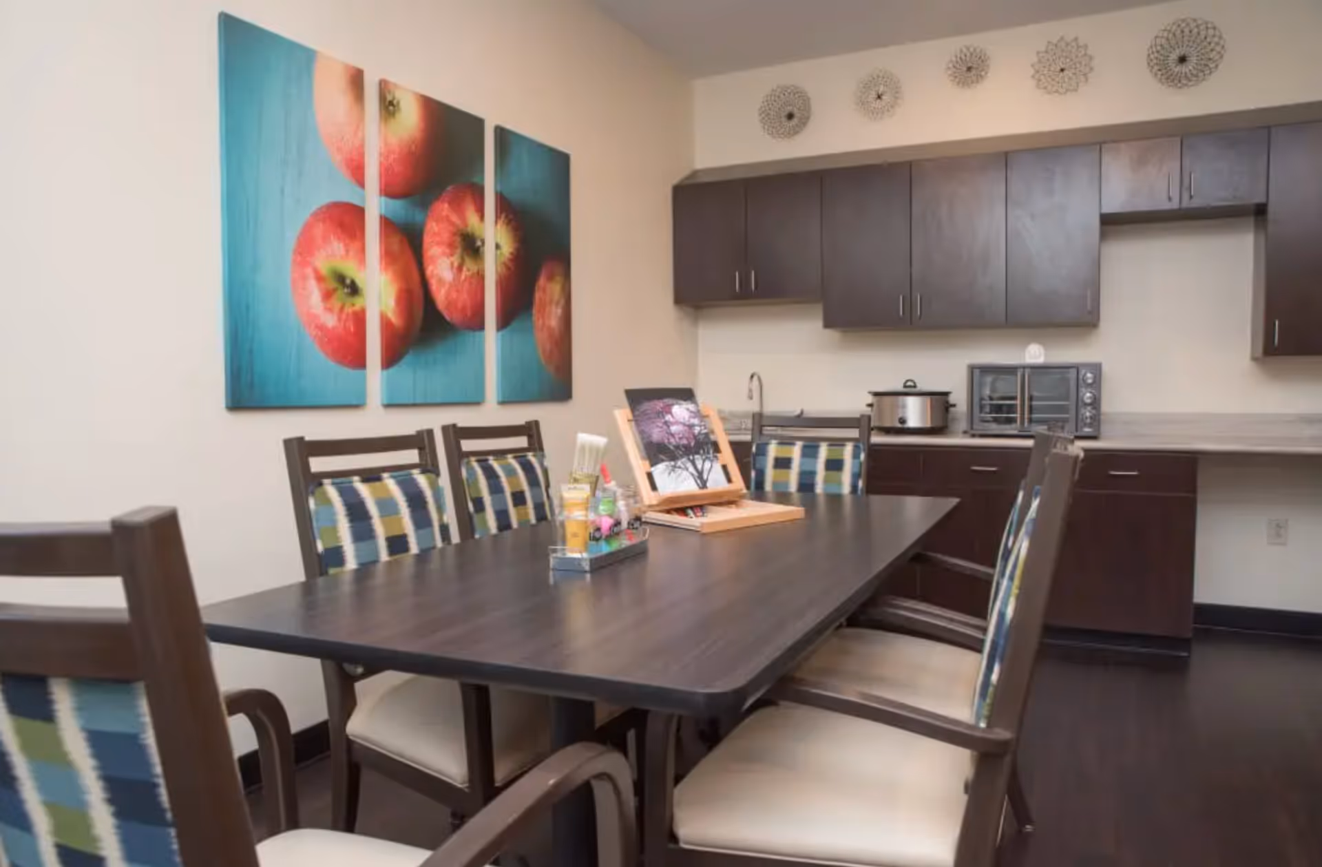 A dining area with a dark wooden table surrounded by six chairs with patterned cushions. On the table, there is a small art easel displaying a painting and a container holding art supplies. Behind the table, there is a kitchenette with dark cabinets, a sink, a slow cooker, and a toaster oven. The wall features a three-panel artwork of red apples on a blue background and decorative circular wall hangings above the cabinets.