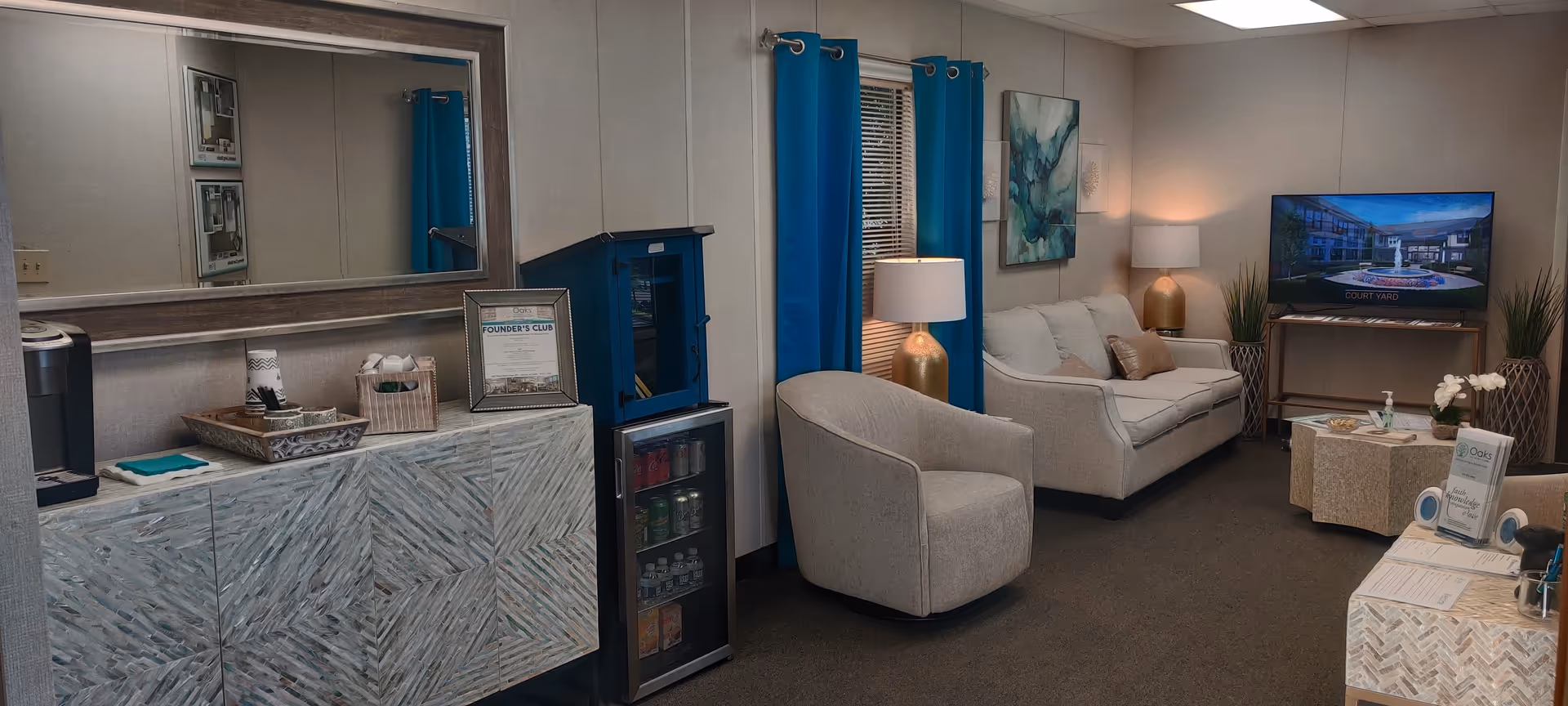 A cozy living room area in a senior living facility with a beige sofa, a matching armchair, a TV on a stand displaying an image of a courtyard, blue curtains on the window, two gold table lamps, decorative plants, and a large mirror above a patterned cabinet with a small refrigerator next to it.