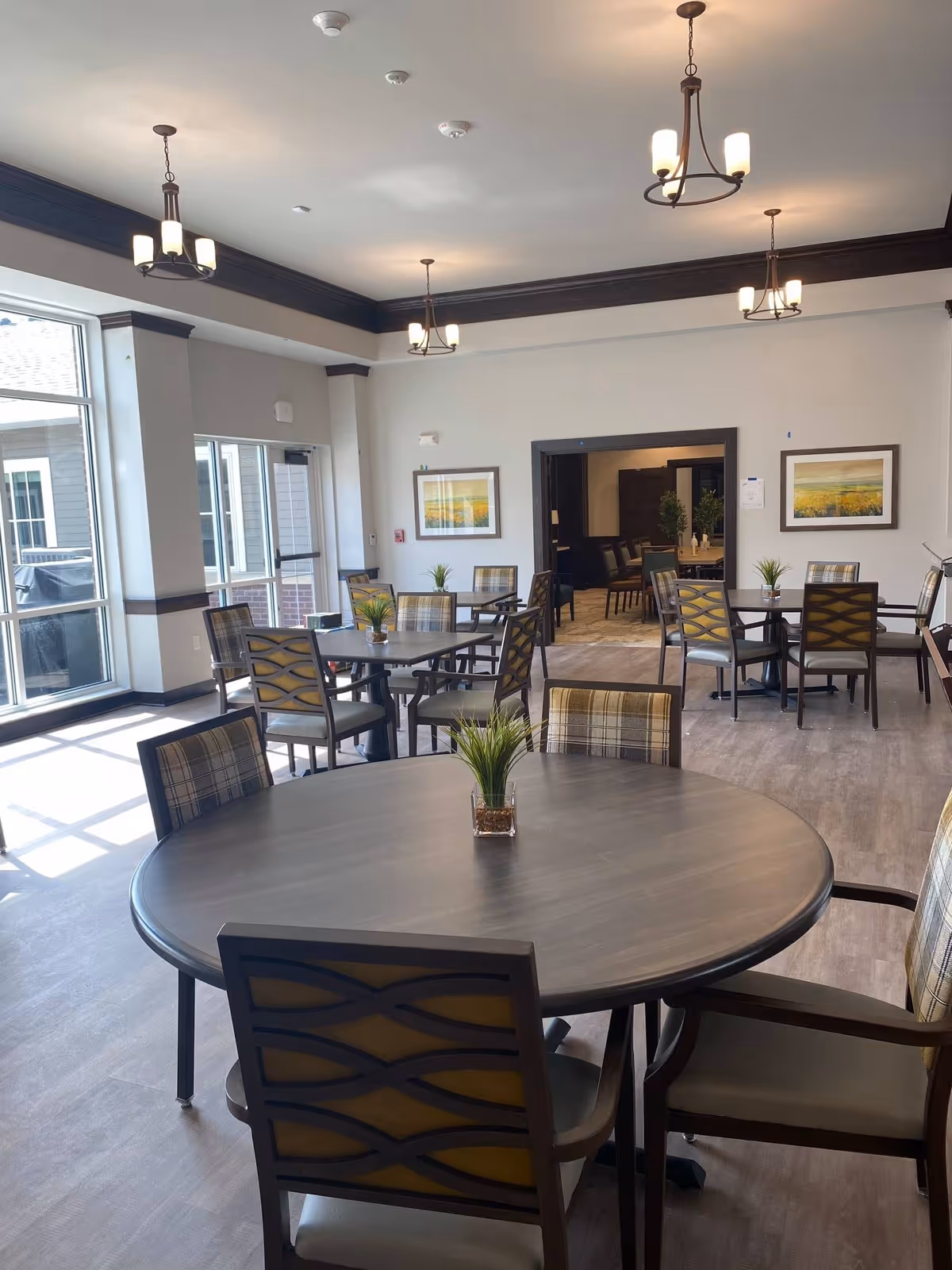 Well-lit communal dining room with round tables, chairs, pendant lights, and large windows.