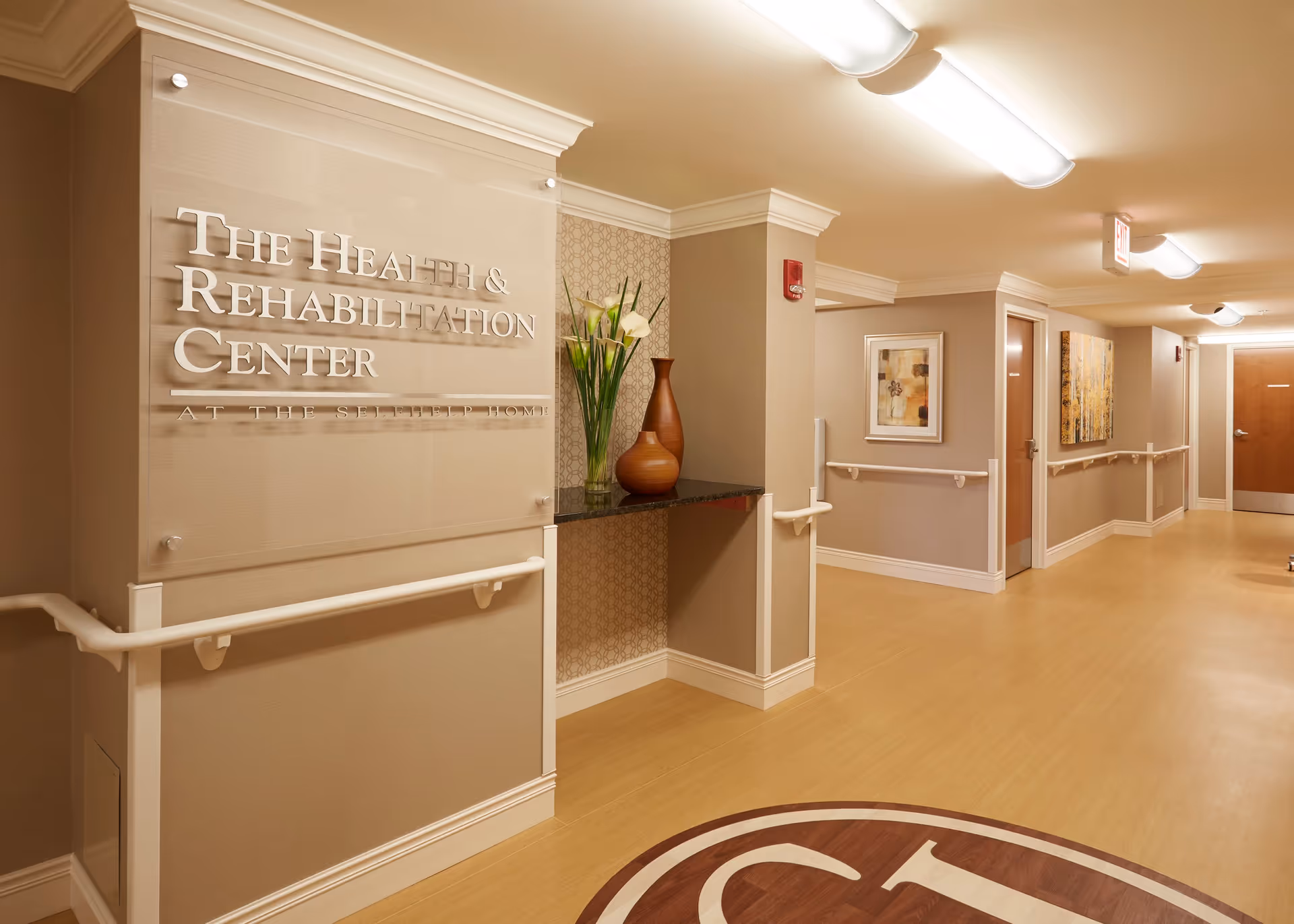 Interior hallway of The Health & Rehabilitation Center at The Selfhelp Home, featuring beige walls with handrails, framed artwork, a decorative shelf with vases and flowers, and overhead fluorescent lighting.