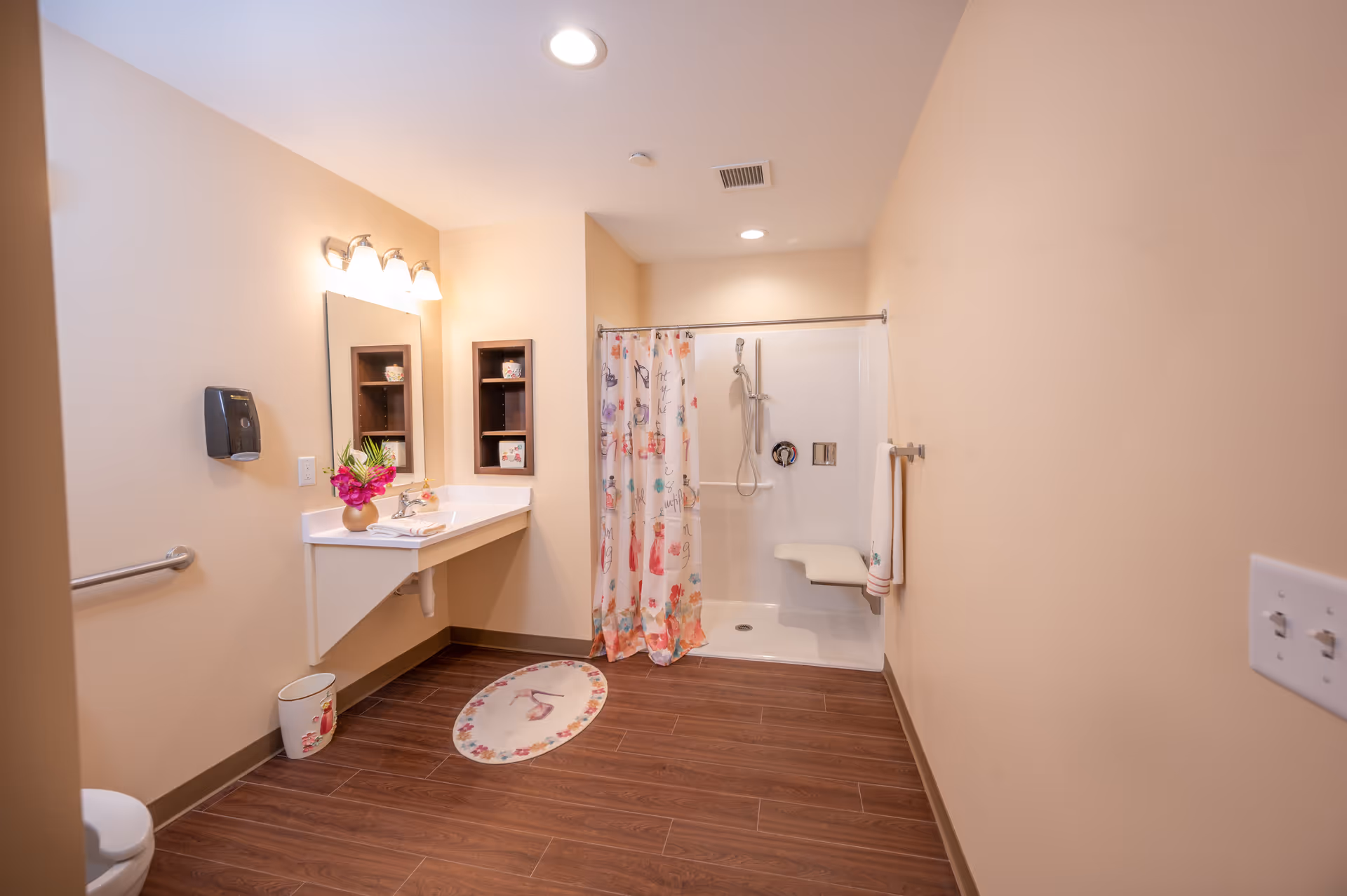 A spacious bathroom with beige walls and wood-look flooring. The room features a white wall-mounted sink with a mirror above it, illuminated by three light fixtures. There is a floral arrangement on the sink counter. To the right, there is a walk-in shower with a floral shower curtain, a handheld showerhead, and a fold-down shower seat. A towel hangs on a bar next to the shower. The bathroom also has a small wall-mounted shelving unit and a wastebasket with a floral design.