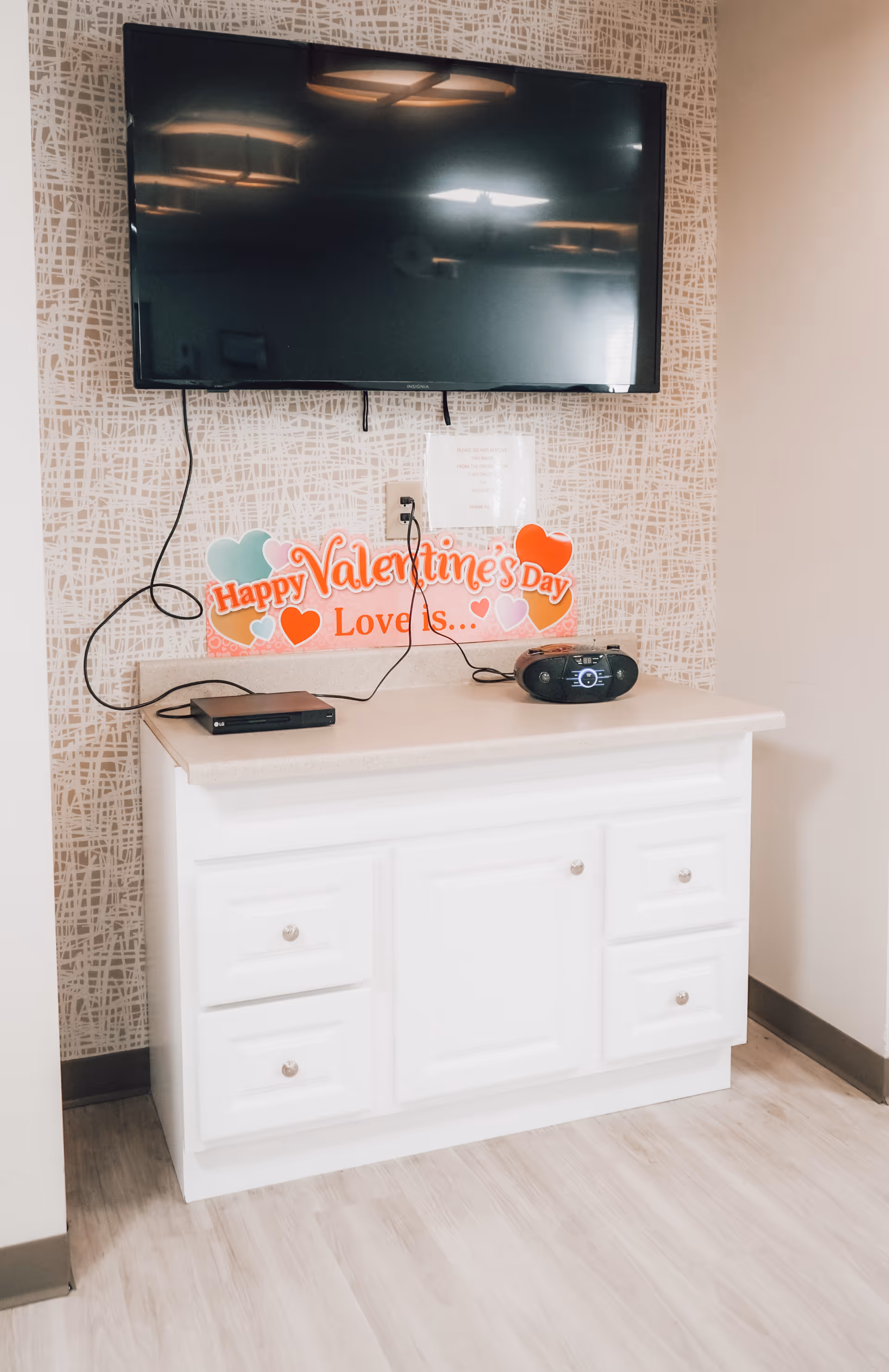 A white cabinet with drawers and a countertop, on which a small black radio and a black electronic device are placed. Above the cabinet, a flat-screen TV is mounted on a patterned wall. A colorful Valentine’s Day decoration with hearts and the text 'Happy Valentine's Day Love is...' is displayed on the wall below the TV.