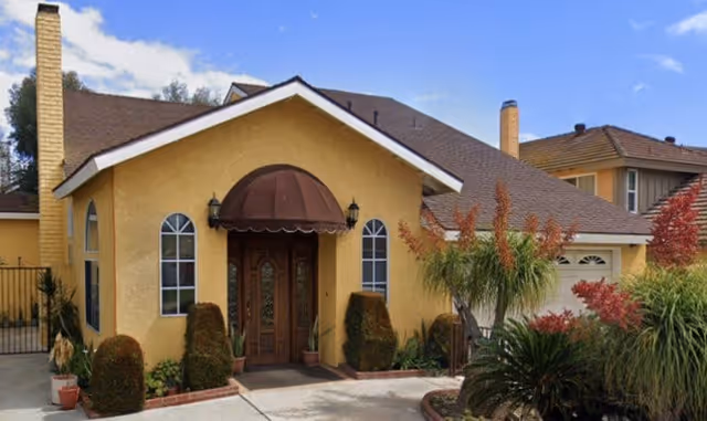 Front exterior of a yellow stucco house with a brown awning over a wooden double door, arched windows, and landscaped shrubs.