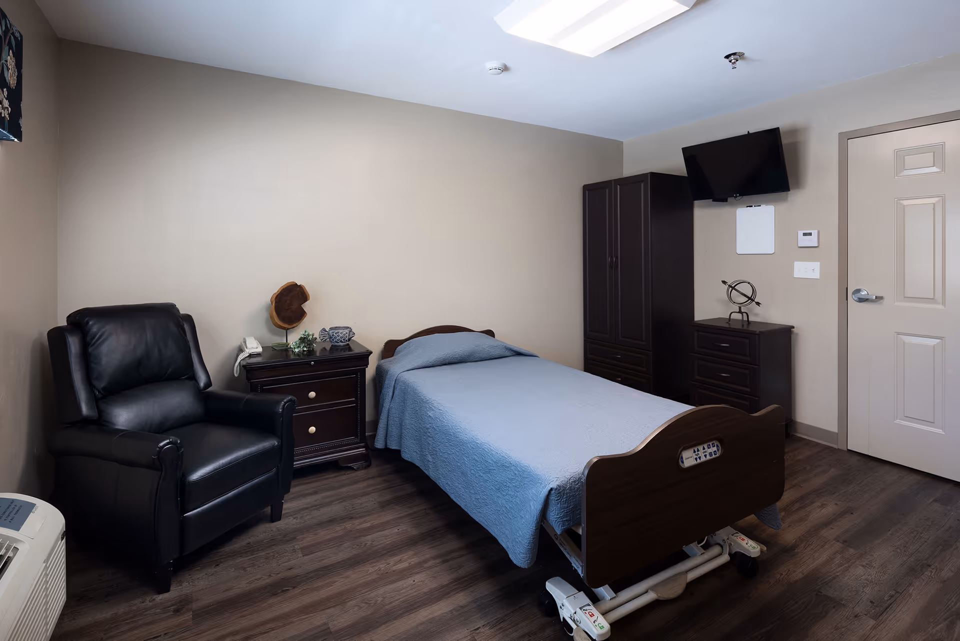 A simple senior-care bedroom with a single adjustable bed, black recliner, nightstand, wardrobe, and wall-mounted TV.