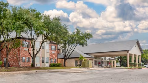 Front entrance of a brick senior living building with a covered porte-cochère, trees, and a partly cloudy sky.
