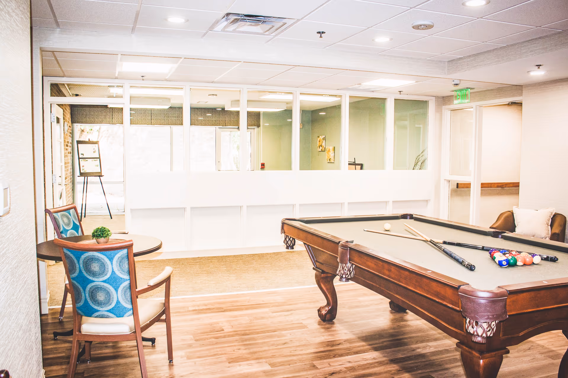 A bright recreational room featuring a pool table with cues and balls arranged on it, a small round table with two chairs upholstered in blue patterned fabric, and a cushioned armchair with a pillow. The room has wood flooring, white walls, and a ceiling with recessed lighting. Large windows and glass doors are visible in the background.