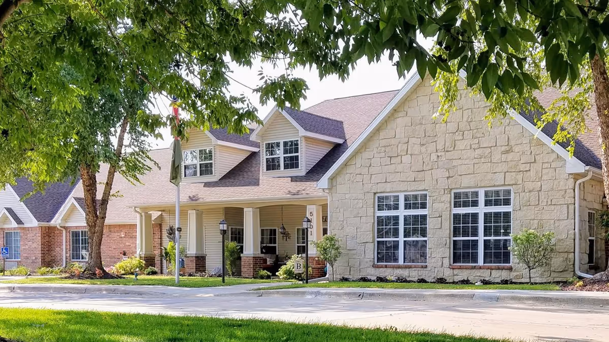 Exterior view of a senior living facility building with a stone and brick facade, large windows, and a covered entrance. The building is surrounded by green trees and a well-maintained lawn, with a driveway in front.
