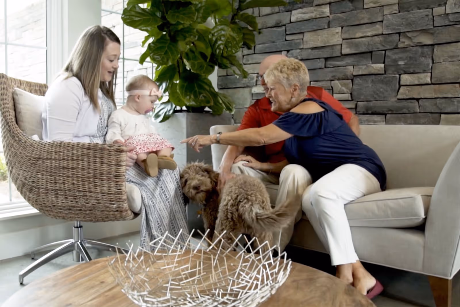 A family scene in a living room with a woman holding a baby sitting on a wicker chair, an elderly woman reaching out to pet two small dogs, and an elderly man sitting on a beige couch against a stone wall. There is a large green plant in the background and a wooden coffee table with a decorative metal bowl in the foreground.