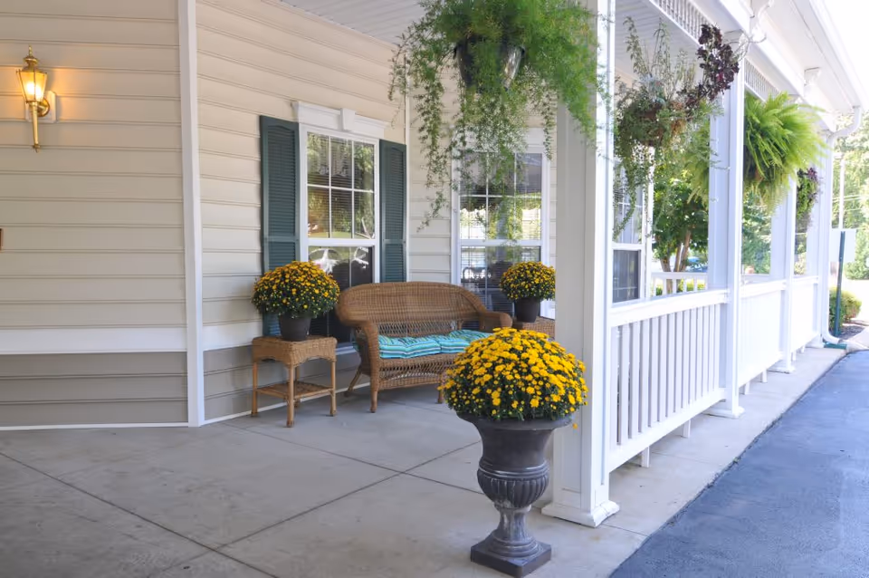 A covered porch area with a wicker bench and a small wicker table, both adorned with yellow potted flowers. Hanging plants are suspended from the porch ceiling, and there are windows with green shutters on the beige siding wall. A lantern-style light fixture is mounted on the wall, and the porch overlooks a paved driveway.