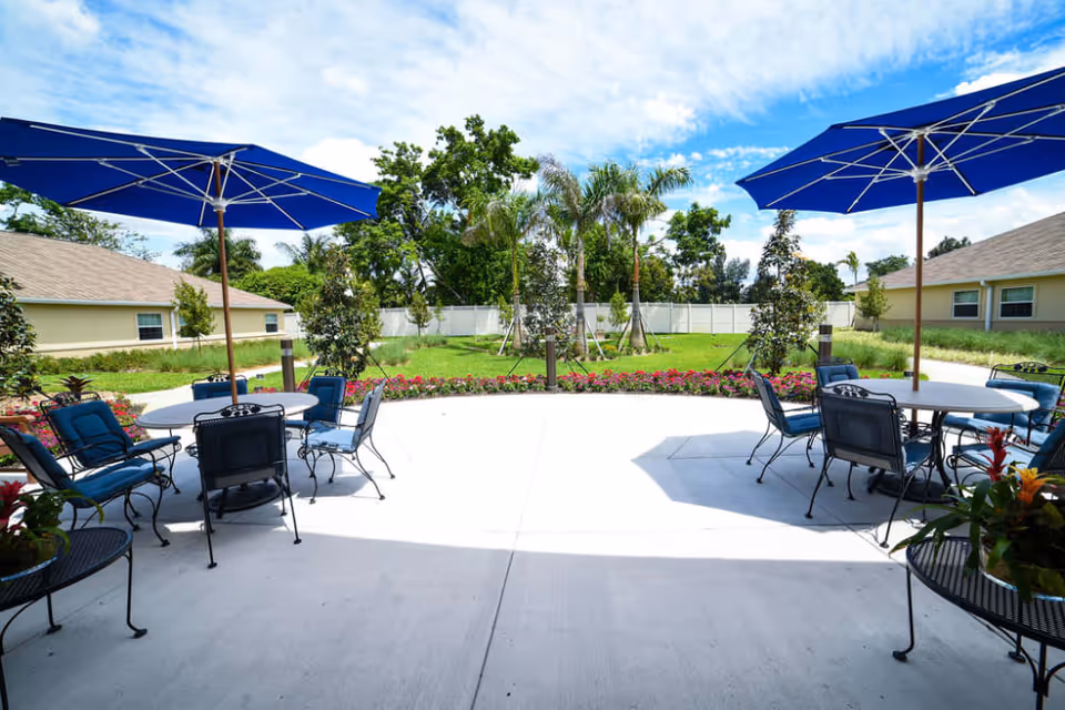 Outdoor patio area with round tables and blue cushioned chairs under large blue umbrellas. The patio overlooks a garden with green grass, colorful flowers, palm trees, and other greenery, surrounded by a white fence and single-story buildings in the background under a partly cloudy sky.
