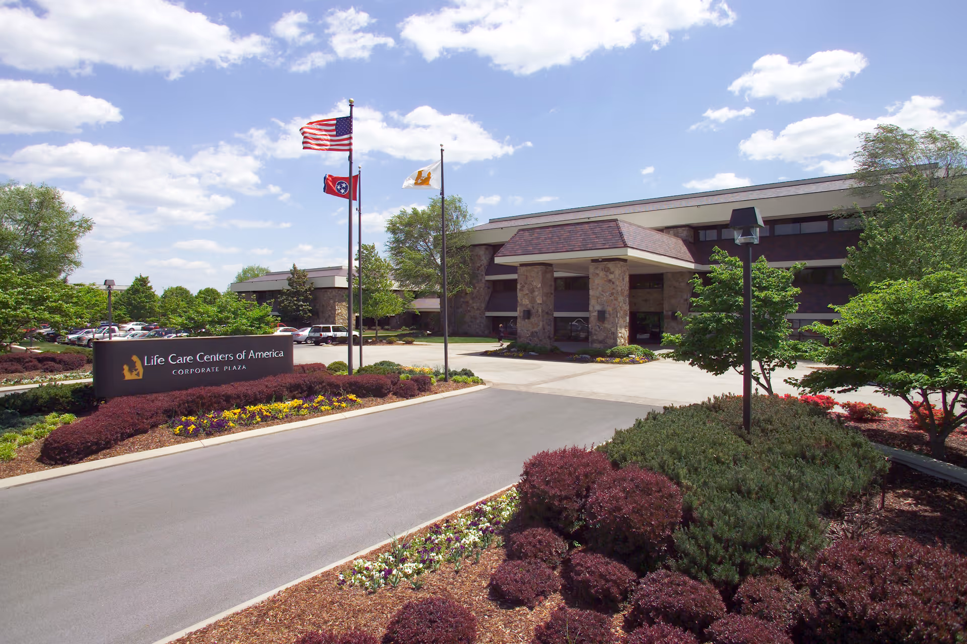 Exterior view of Life Care Centers of America Corporate Plaza building with a driveway, landscaped bushes, flowers, and three flagpoles displaying the American flag, Tennessee state flag, and another flag under a partly cloudy sky.