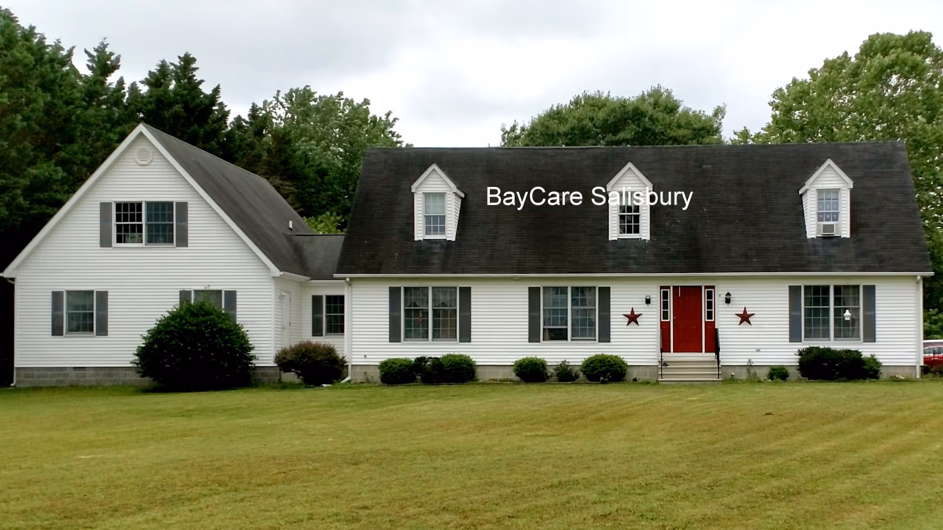 Front exterior view of a white two-story house with a dark roof, red double front doors, and multiple windows. The house is surrounded by green grass and trees in the background. The text 'BayCare Salisbury' is overlaid on the roof area.