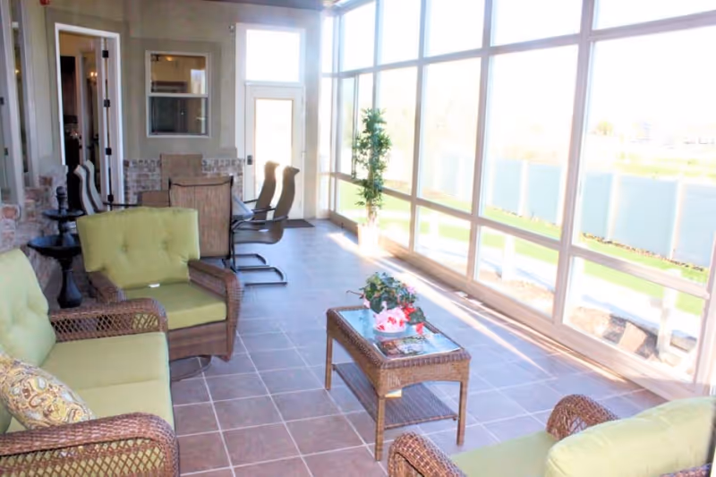 Sunlit sunroom-style sitting area with wicker sofas and chairs, a coffee table, and floor-to-ceiling windows.