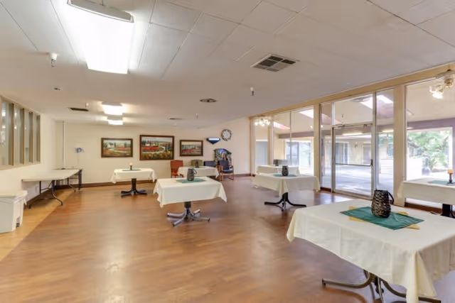 Spacious dining/activity room with several small tables covered in white tablecloths and glass doors leading to an outdoor patio.