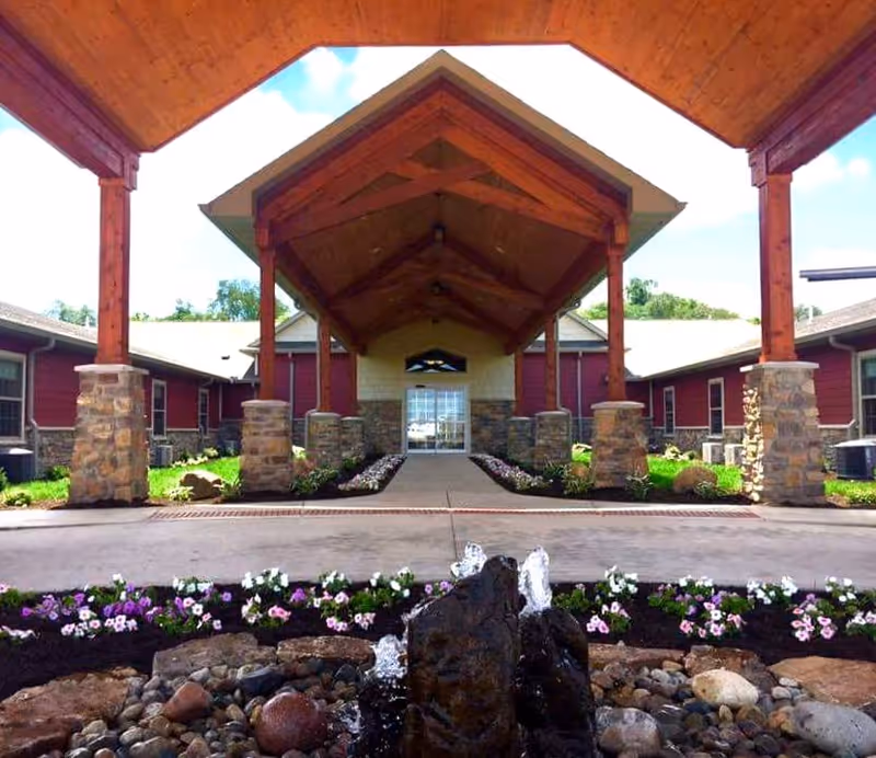 Entrance of Copper Creek Cottages Memory Care of Mattoon featuring a covered walkway with wooden beams and stone pillars, landscaped flower beds, and a small rock water fountain in the foreground.