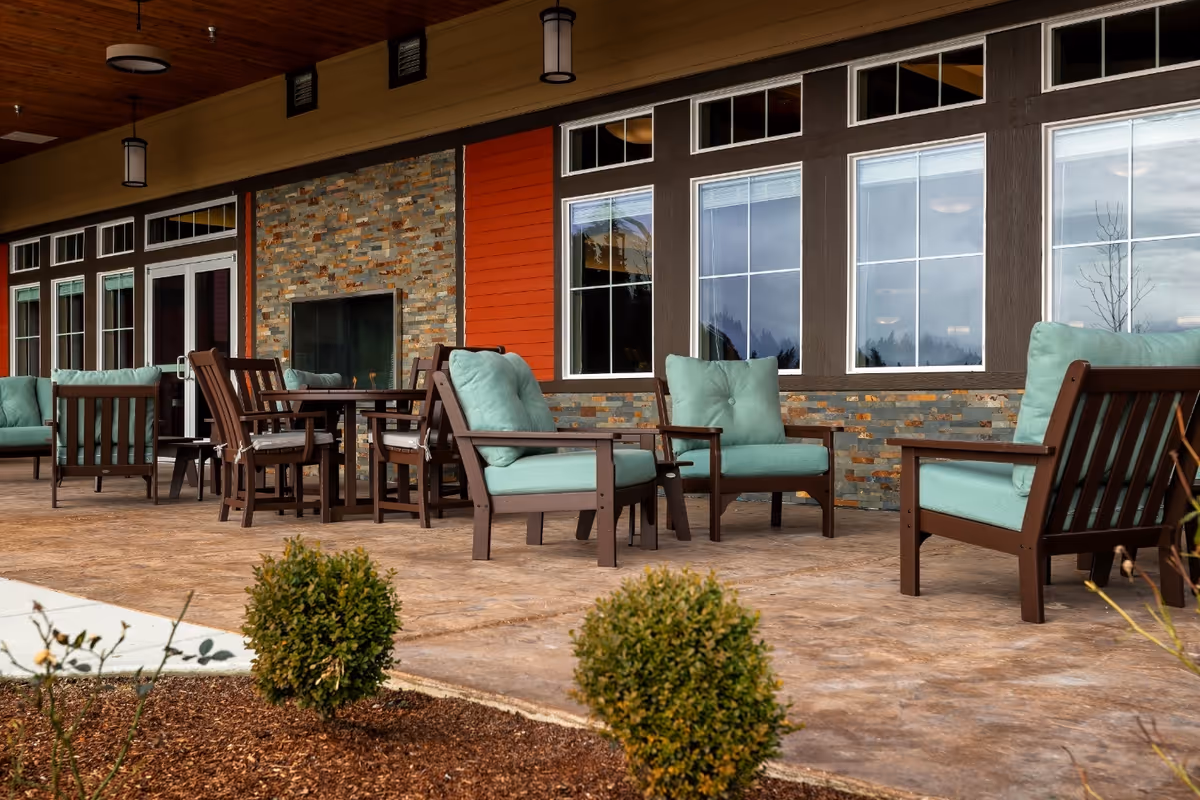 Outdoor patio area with cushioned wooden chairs and tables arranged on a stone floor in front of a building with large windows and a stone and wood exterior wall.