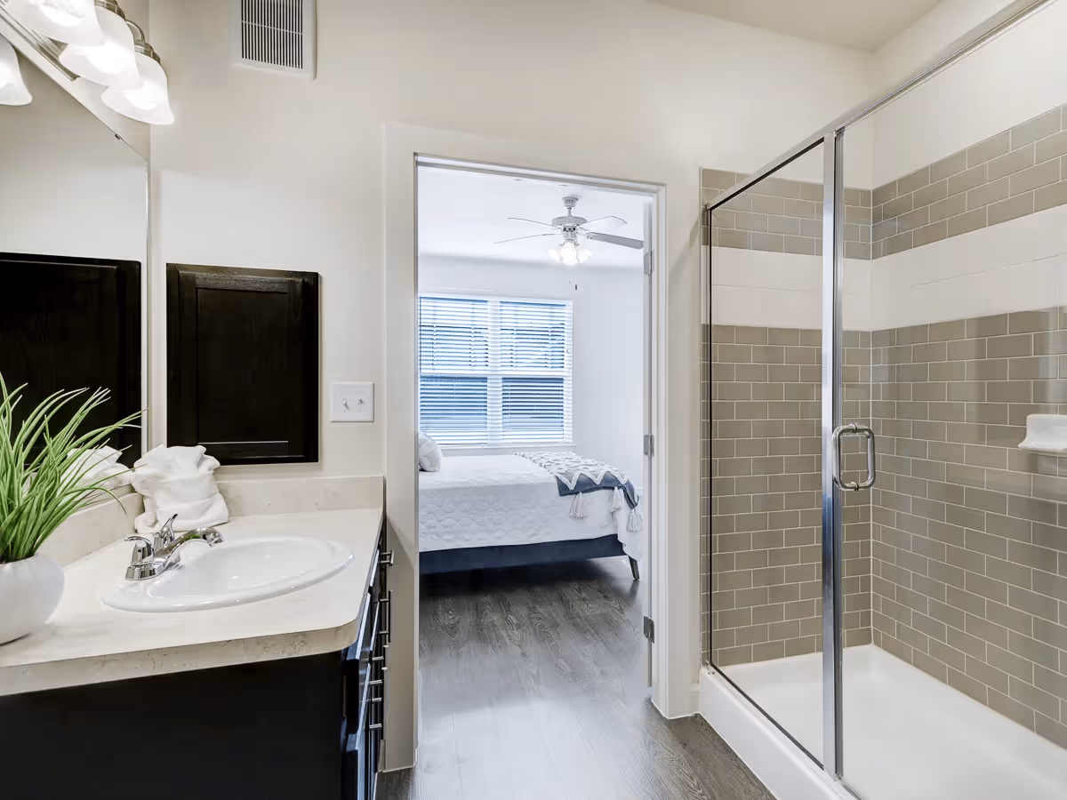 Bathroom with a white sink and countertop, a potted plant, and a mirror on the left. To the right is a glass-enclosed shower with beige and white tiled walls. Through an open doorway, a bedroom with a bed, white bedding, and a ceiling fan is visible.