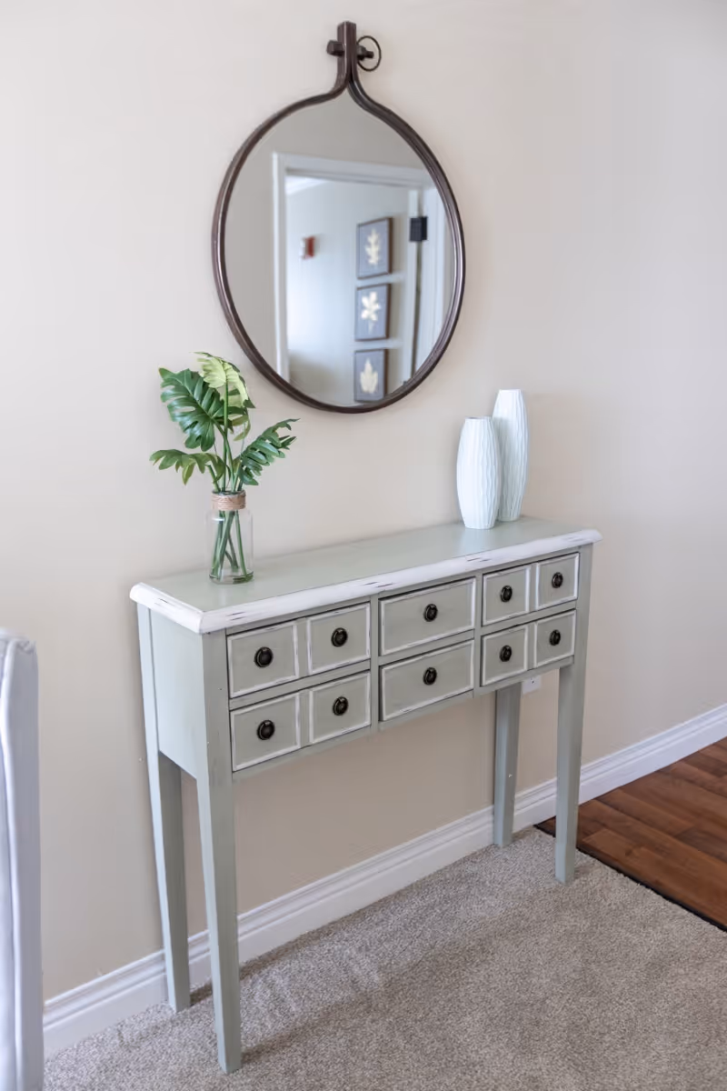 A narrow console table with multiple small drawers stands against a beige wall. On top of the table are two white decorative vases and a glass jar with green leafy plants. Above the table hangs a round mirror with a dark frame reflecting part of the opposite wall with framed artwork.