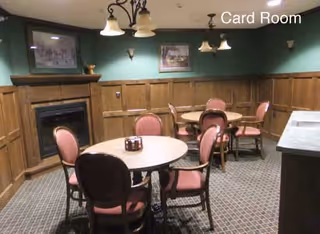 A cozy card room with round tables and pink cushioned chairs arranged around them. The room features wood-paneled walls, a fireplace, framed artwork, and soft lighting from ceiling fixtures. The carpet has a patterned design and the words 'Card Room' are visible on the upper right corner of the image.
