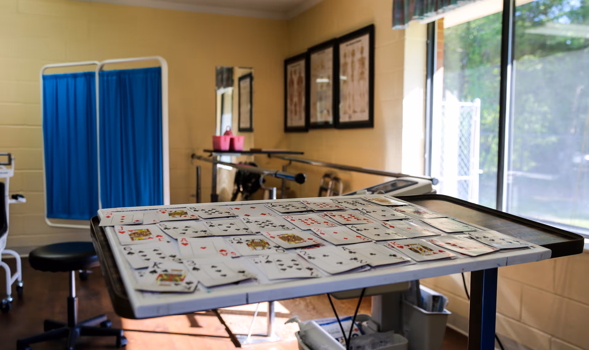 A therapy or rehabilitation room with a table covered in playing cards, a blue privacy screen, a stool, parallel bars for walking exercises, anatomical charts on the wall, and a large window letting in natural light.