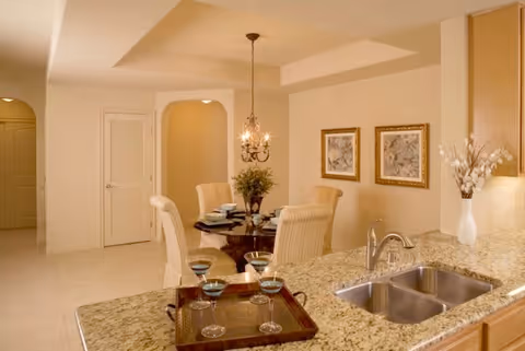 View of a dining area and kitchen counter in a senior living facility. The dining area features a round glass table with four upholstered chairs, a chandelier hanging above, and two framed pictures on the wall. The kitchen counter has a granite surface with a double sink and a tray holding four wine glasses. A vase with flowers is placed near the sink.