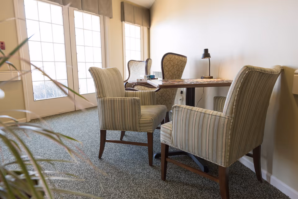 Small seating area with upholstered armchairs around a wooden table by large windows in a senior living facility.