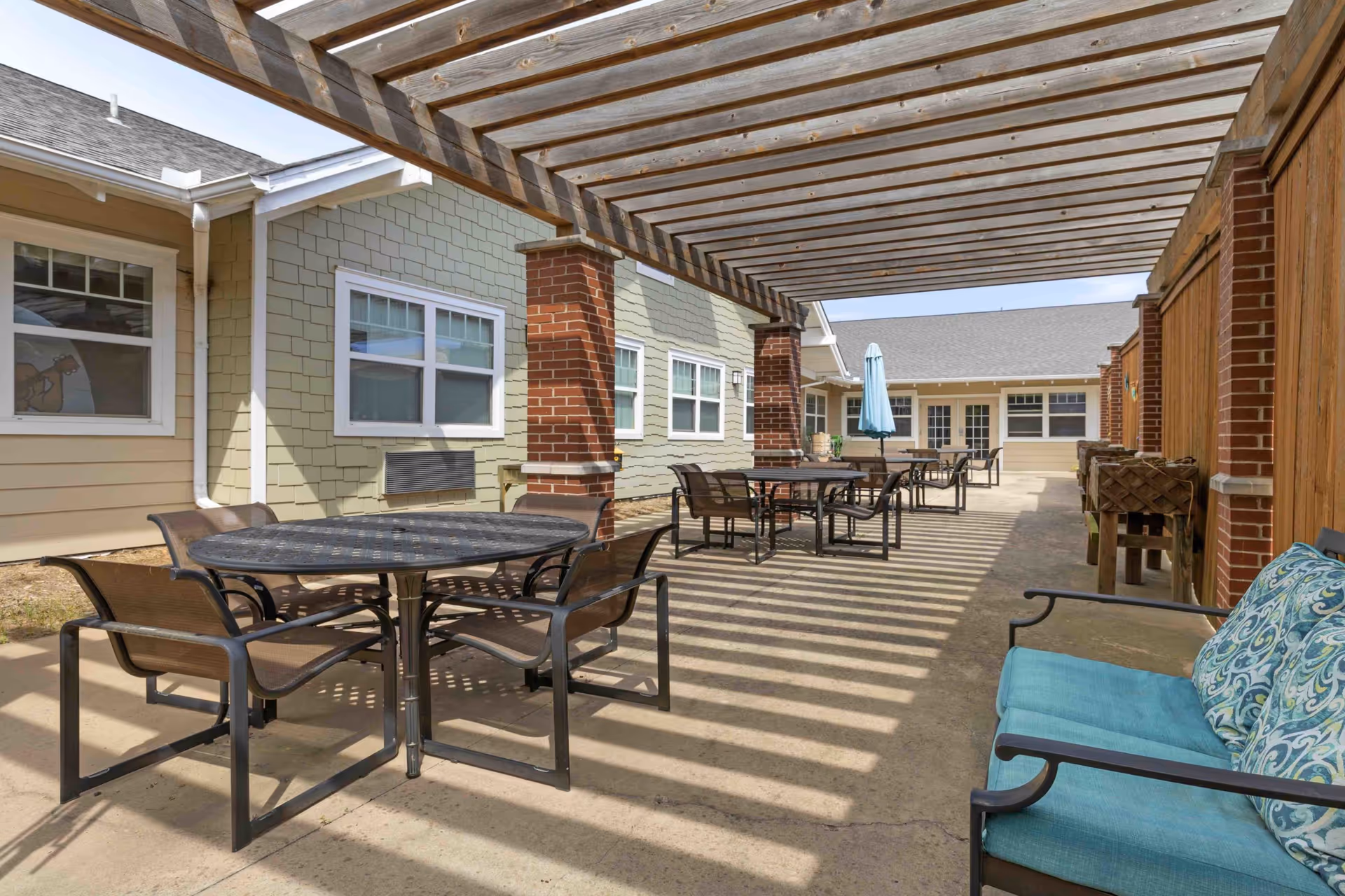 Outdoor patio area at Aberdeen Memory Care of Tulsa with multiple round tables and chairs under a wooden pergola. There is a cushioned bench with patterned pillows on the right side and a building with windows and doors in the background.