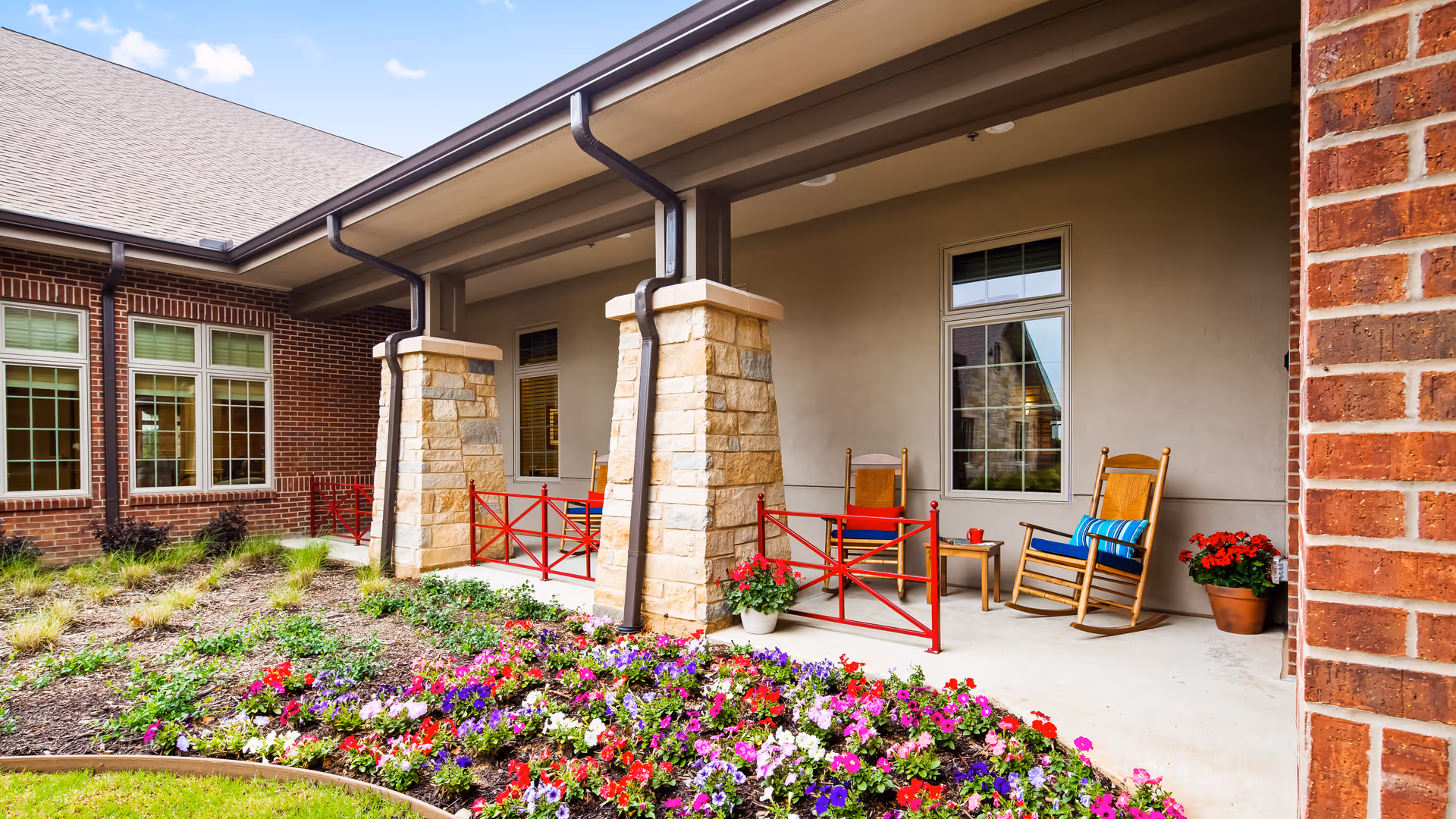 Covered outdoor patio with rocking chairs, potted plants, and a colorful flowerbed in front of a brick building.