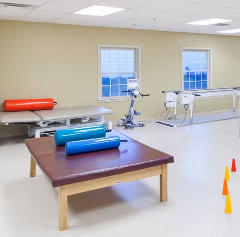 A physical therapy room with exercise equipment including a padded treatment table with red and blue cylindrical foam rollers, a stationary exercise bike, parallel bars for walking exercises, and several small orange and yellow cones on the floor. The room has beige walls and two windows with white frames.