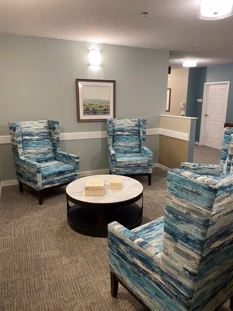 A cozy seating area with four blue patterned armchairs arranged around a round coffee table with two decorative boxes on it. The walls are painted light gray with white trim, and a framed landscape painting hangs on the wall. The space is softly lit by a wall-mounted light fixture and ceiling lights. A hallway with doors is visible in the background.