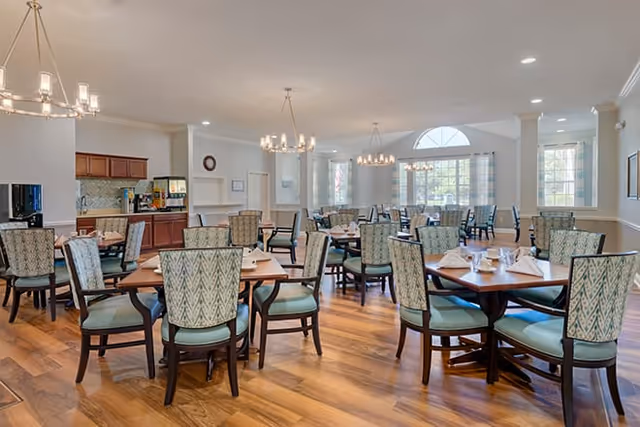 A spacious dining room with multiple wooden tables and cushioned chairs arranged neatly. Each table is set with white napkins and cups. The room has large windows allowing natural light to fill the space, and there are chandeliers hanging from the ceiling. The floor is wooden, and there is a kitchenette area with cabinets and a coffee machine in the background.