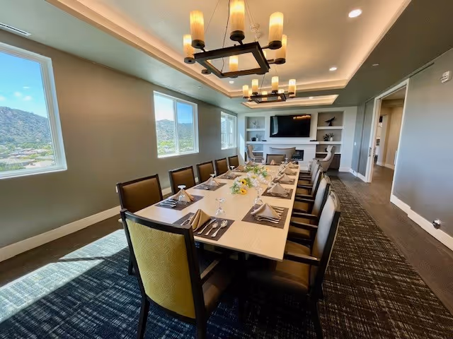 A well-lit dining room with a long rectangular table set for a meal, featuring neatly arranged plates, napkins, and silverware. The room has large windows showing a view of hills outside, modern chandeliers hanging from the ceiling, and a fireplace with a TV above it in the background.