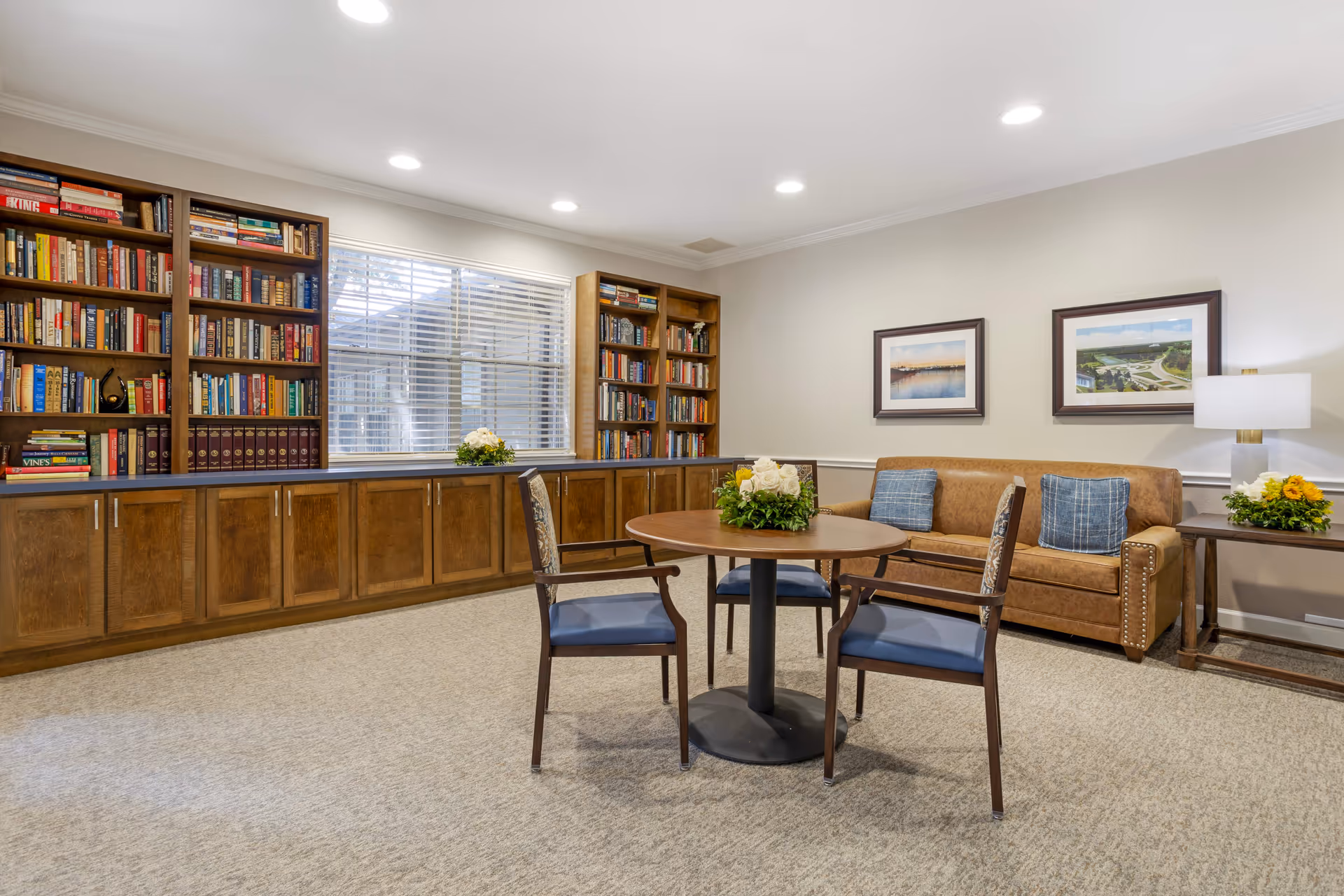 A cozy senior living common room with a round wooden table surrounded by three chairs with blue cushions. Behind the table is a brown leather sofa with two blue pillows. On the right side, there is a wooden side table with a lamp and a flower arrangement. The back wall features two framed pictures. On the left side, there are built-in wooden bookshelves filled with books beneath a window with white blinds. The room is carpeted and well-lit with recessed ceiling lights.