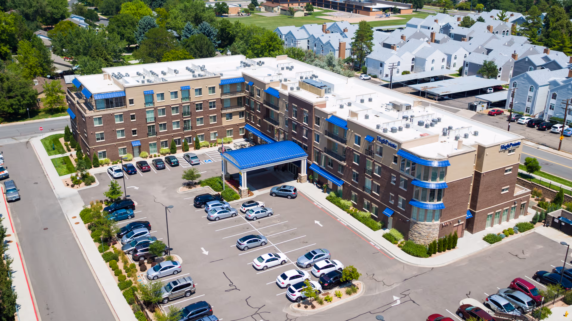Aerial view of HighPointe Assisted Living & Memory Care building with a large parking lot filled with cars surrounding the facility. The building has multiple stories with blue awnings and a covered entrance with a blue roof. Residential houses and trees are visible in the background.