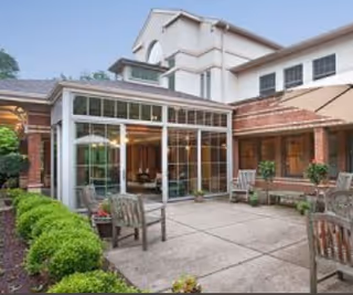 Outdoor patio area at The Pines of Mount Lebanon featuring a paved courtyard with wooden chairs and small tables, surrounded by green shrubs and plants. The building exterior includes large windows and a covered walkway.