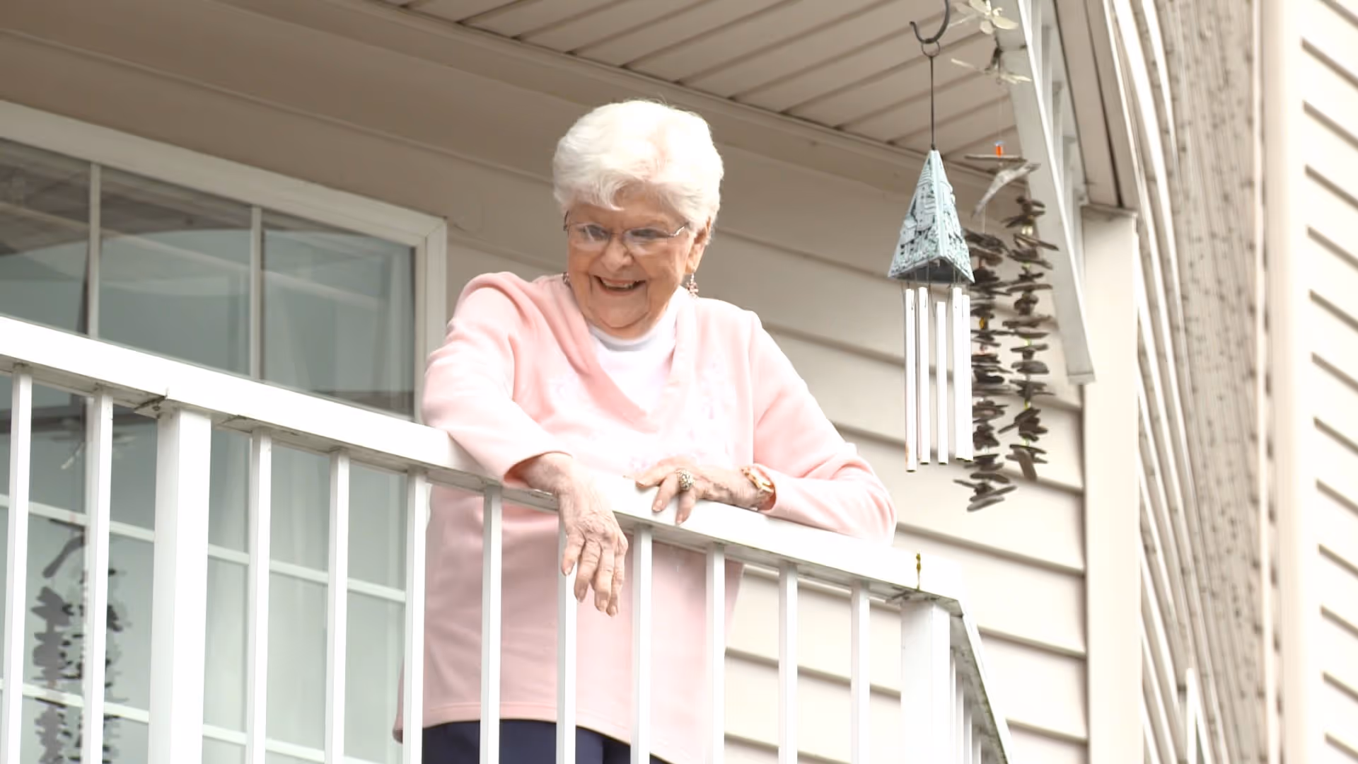 An elderly woman with white hair and glasses wearing a pink sweater stands on a balcony railing outside a building, smiling and looking down. A wind chime hangs nearby.