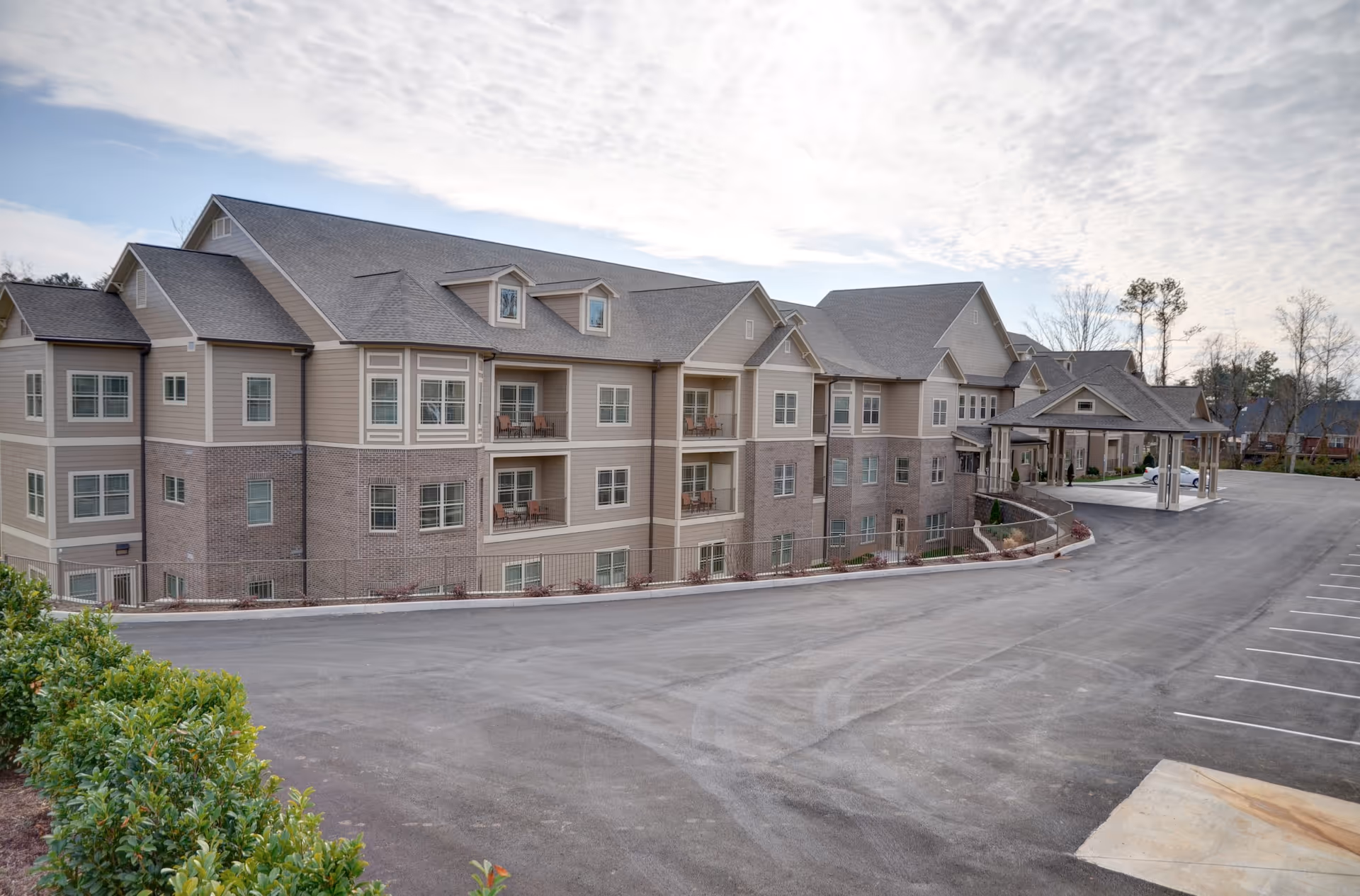 Exterior view of a large, multi-story senior living facility building with beige siding and brick accents, featuring balconies and a covered entrance. The building is surrounded by a paved driveway and parking area, with some greenery in the foreground and trees in the background under a partly cloudy sky.