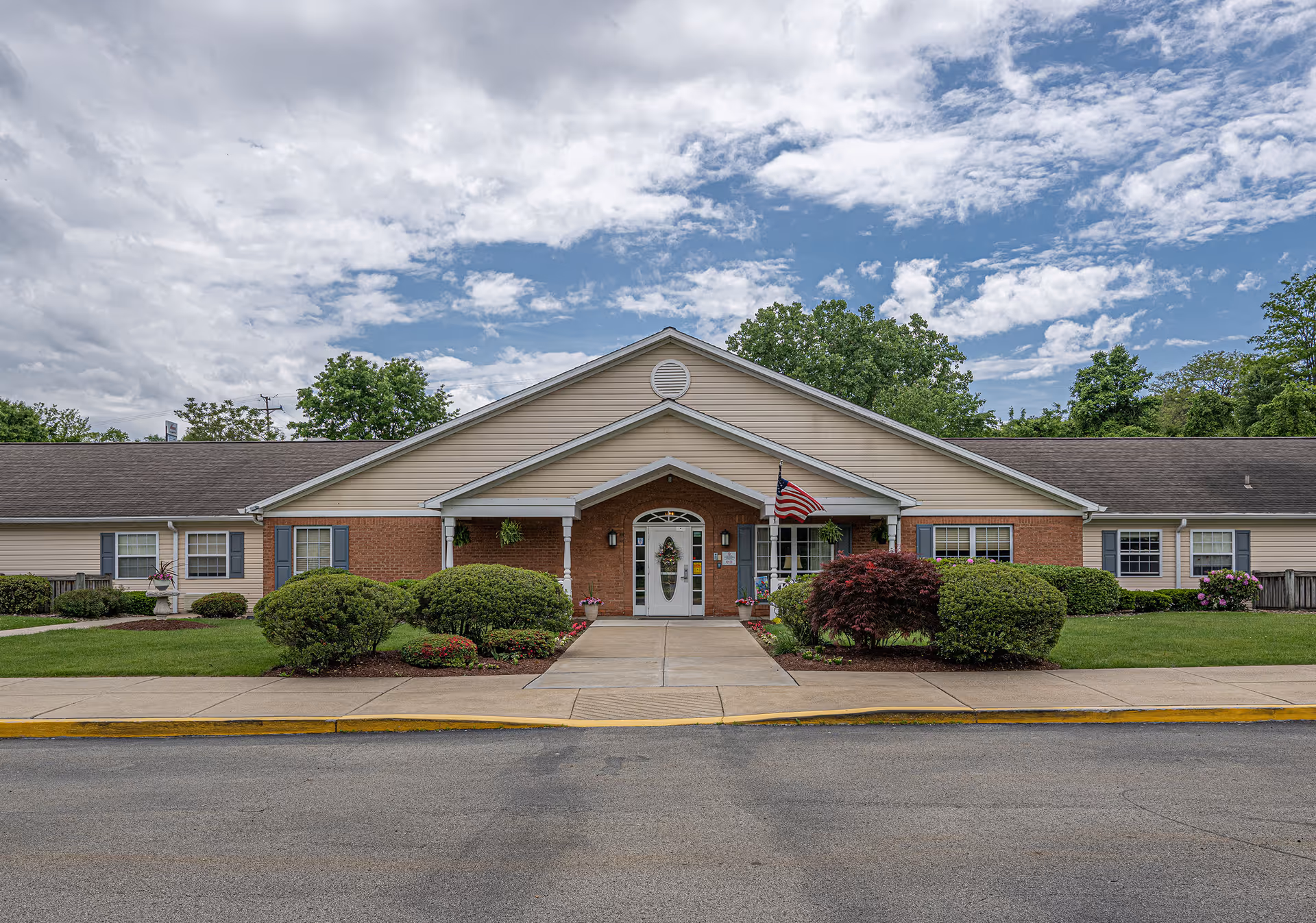 Front exterior view of a single-story building with a peaked roof, beige siding, and red brick accents. The entrance has a covered porch with white columns, an American flag, and well-maintained bushes and greenery along the walkway. The sky is partly cloudy.