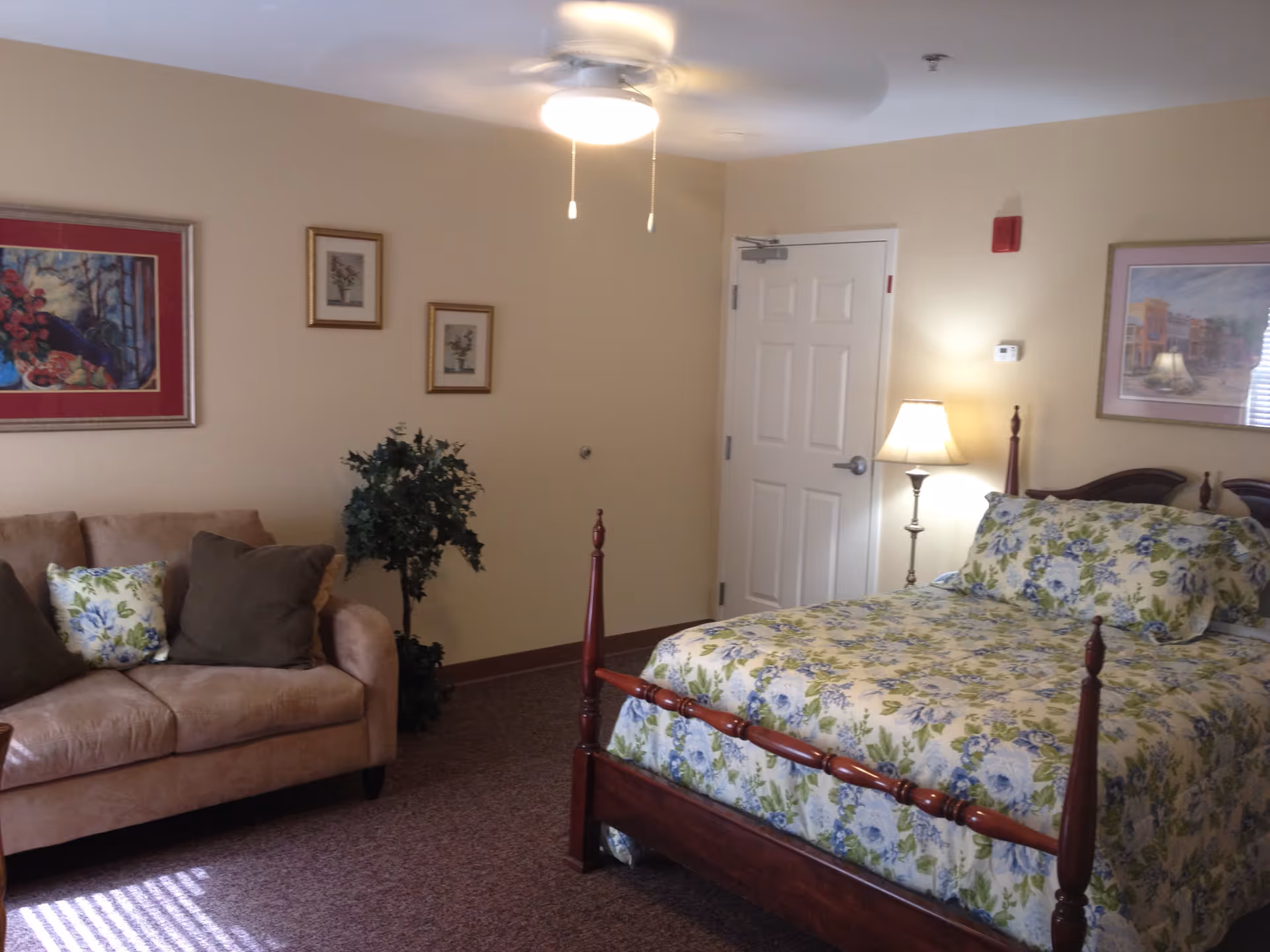 A cozy bedroom with a wooden four-poster bed dressed in floral bedding, a beige couch with decorative pillows, a floor lamp, a ceiling fan with lights, framed artwork on the walls, and a potted plant in the corner.